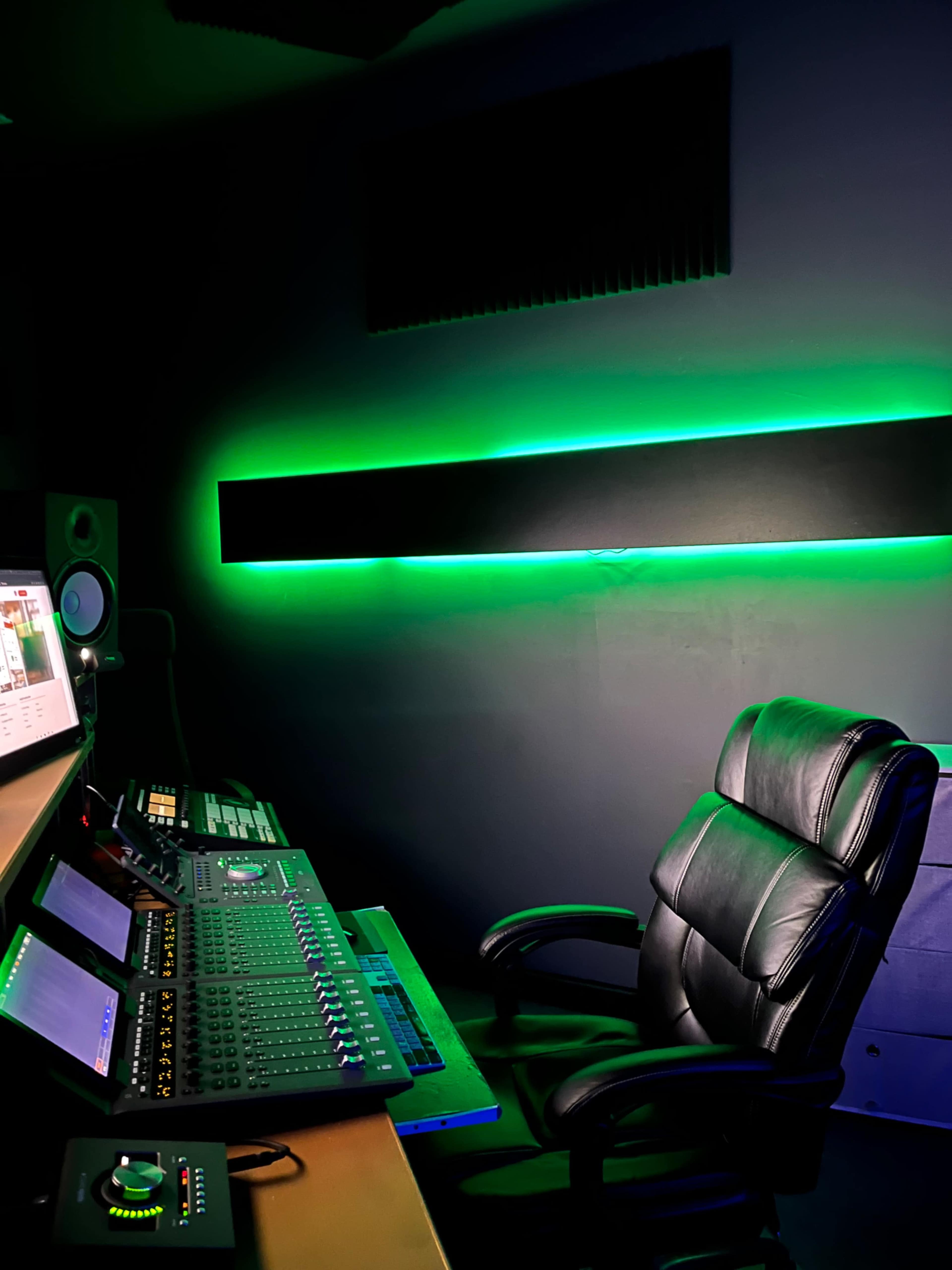A dimly lit music studio featuring a mixing console, computer monitors, and an ergonomic black chair illuminated by green LED lights.
