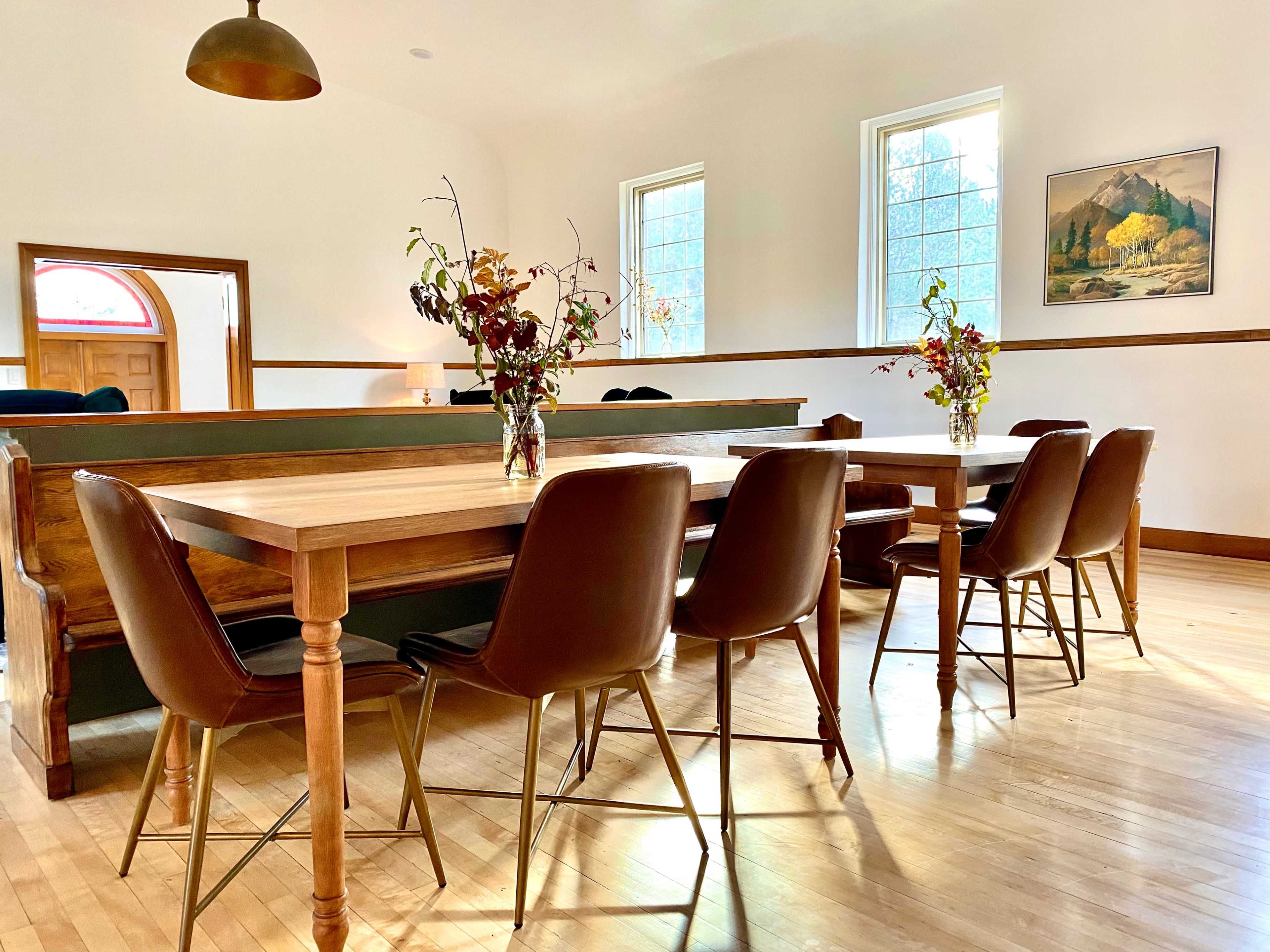 A bright room features two wooden tables surrounded by brown chairs, with vases of flowers on each table and large windows allowing natural light.