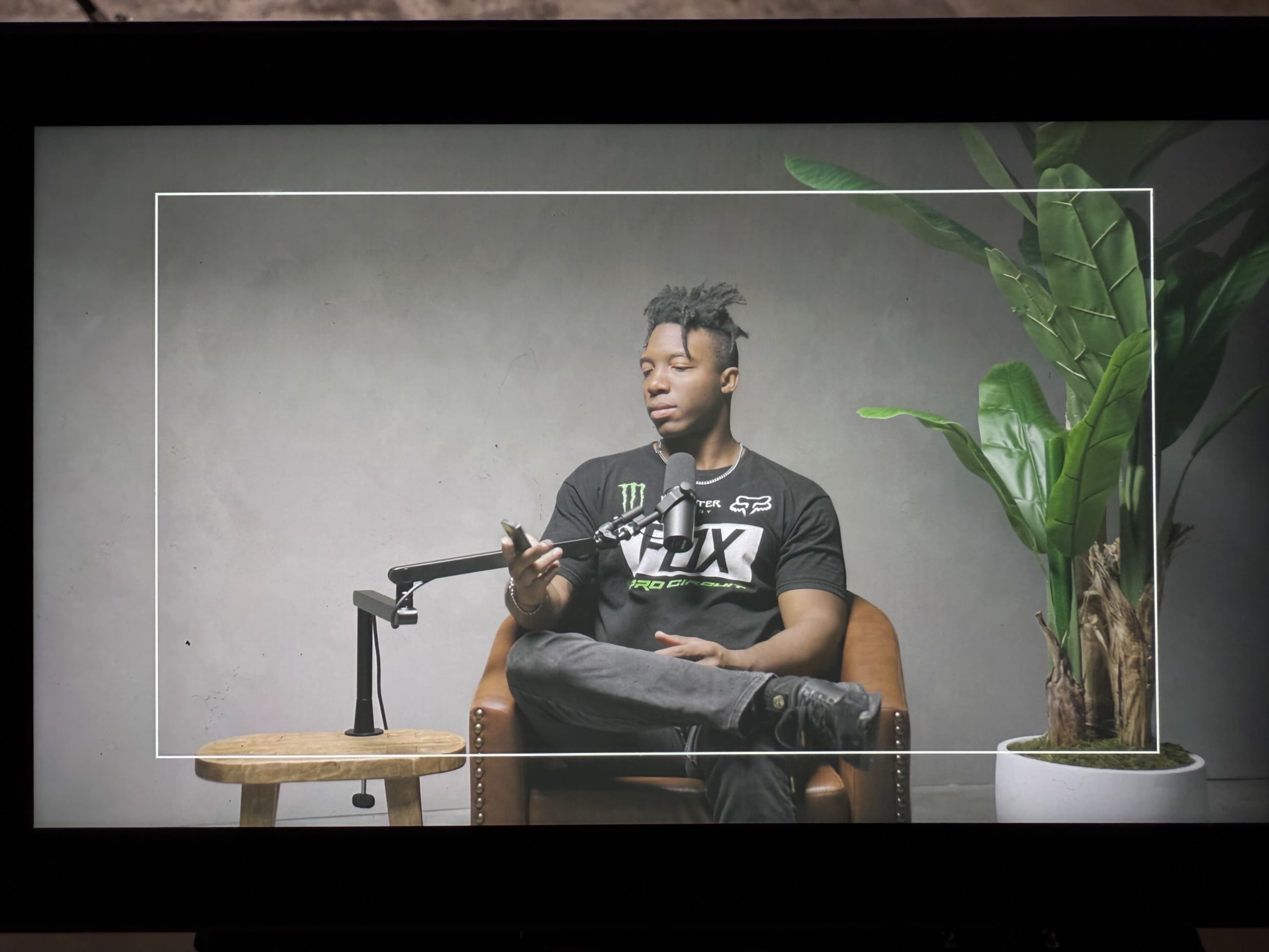 A person sits in a chair holding a phone, with a plant and a stool nearby, against a plain background.