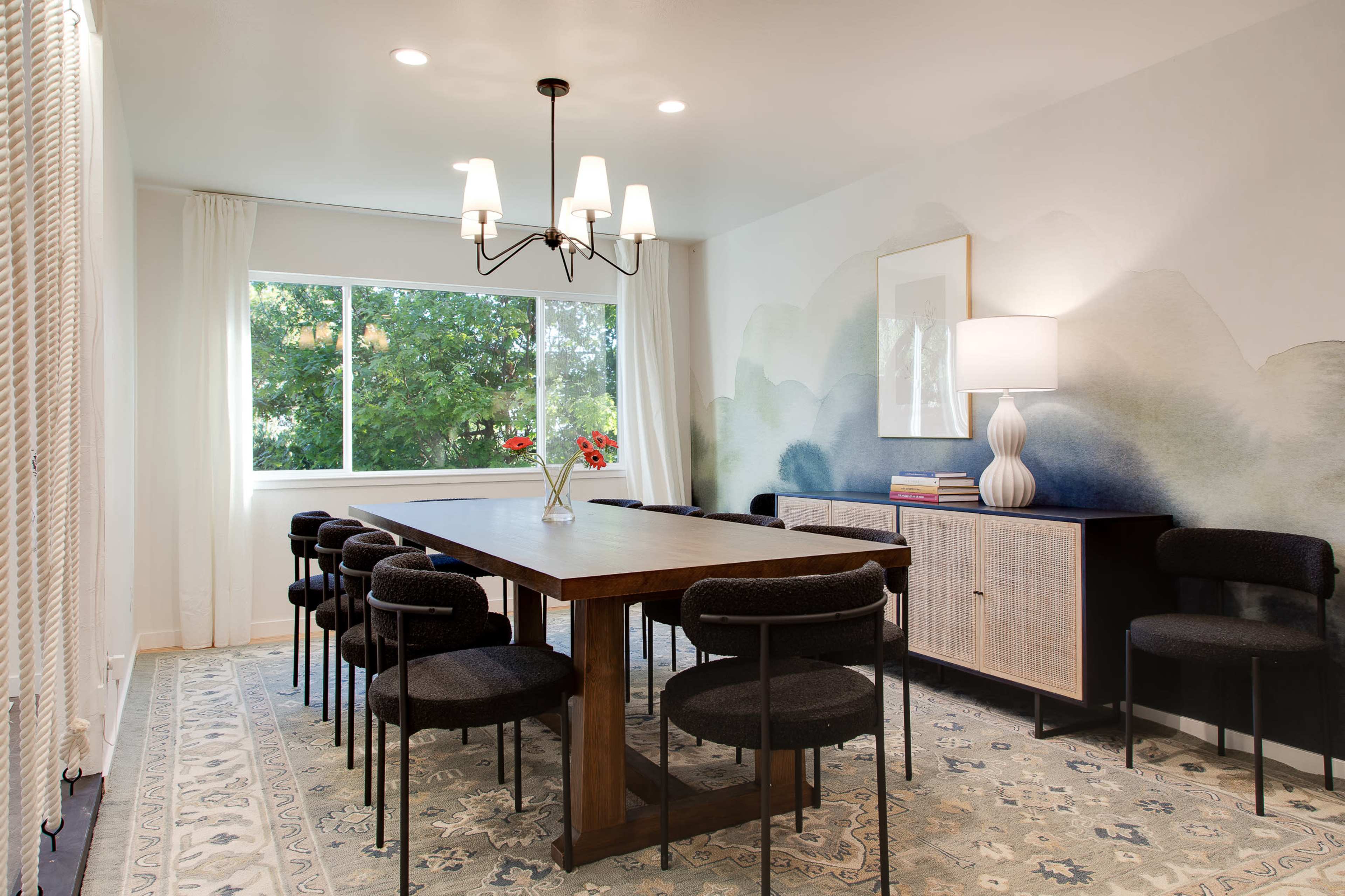 A dining room features a long wooden table surrounded by black chairs, with a large window providing natural light and a mural of soft colors on the wall.