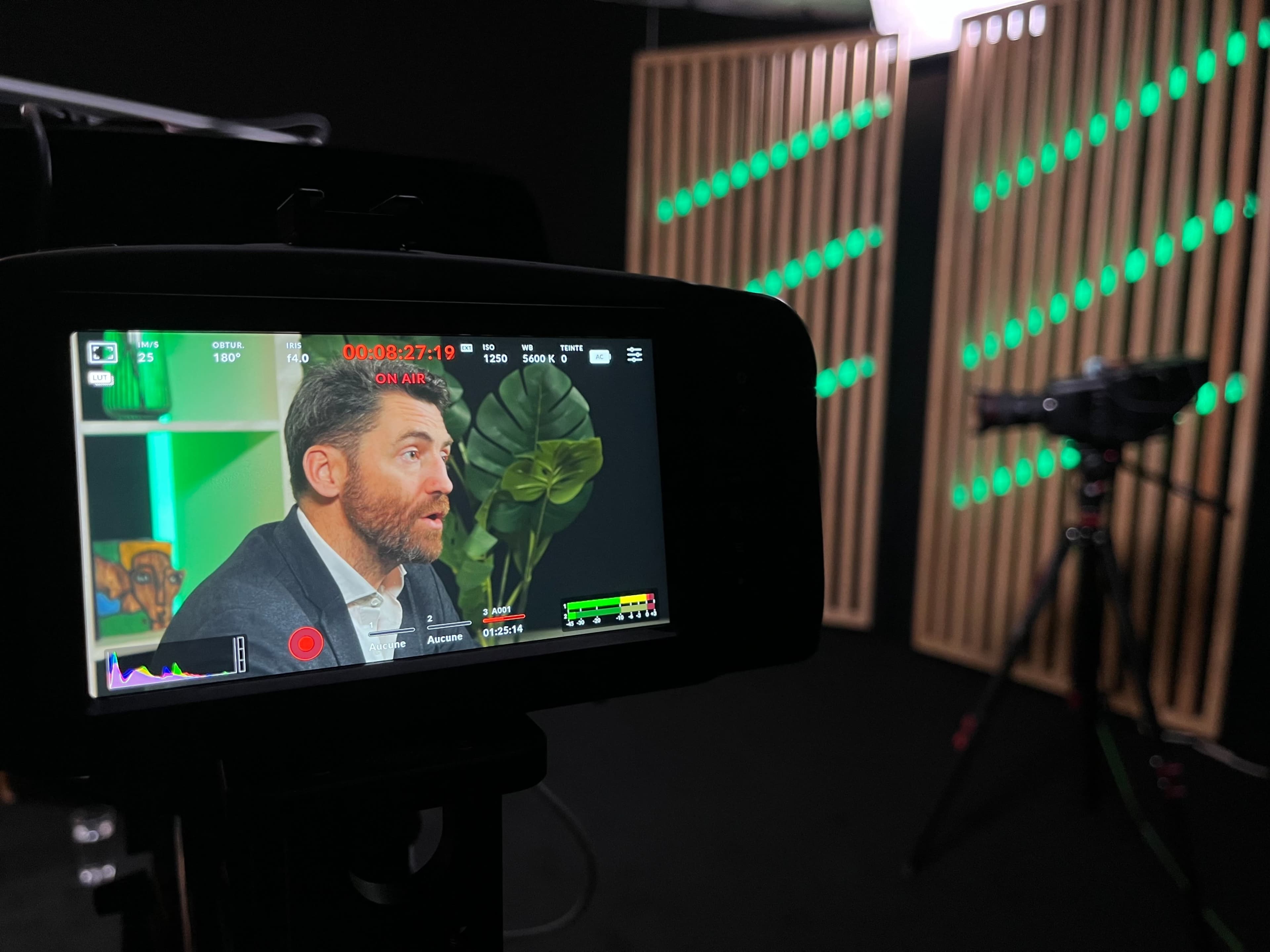 A camera display shows a man speaking in a studio with green lighting and wooden backdrop panels.