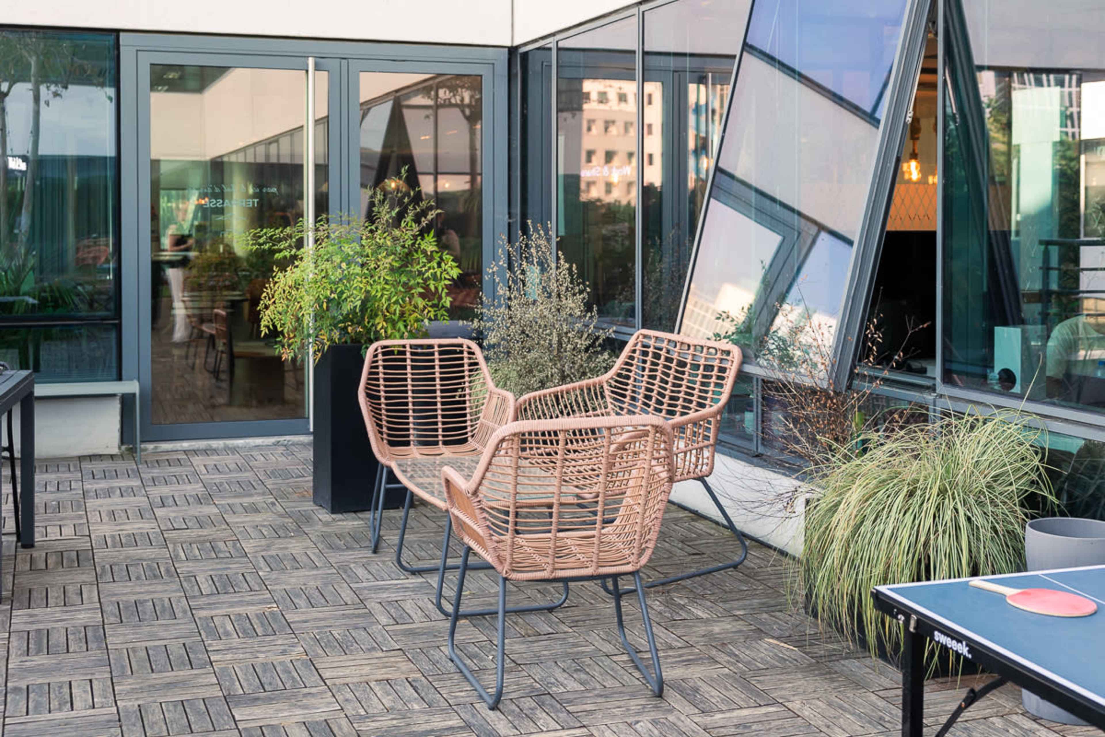 The image shows a modern outdoor patio with two stylish woven chairs, a wooden floor, and large glass windows reflecting the surroundings.