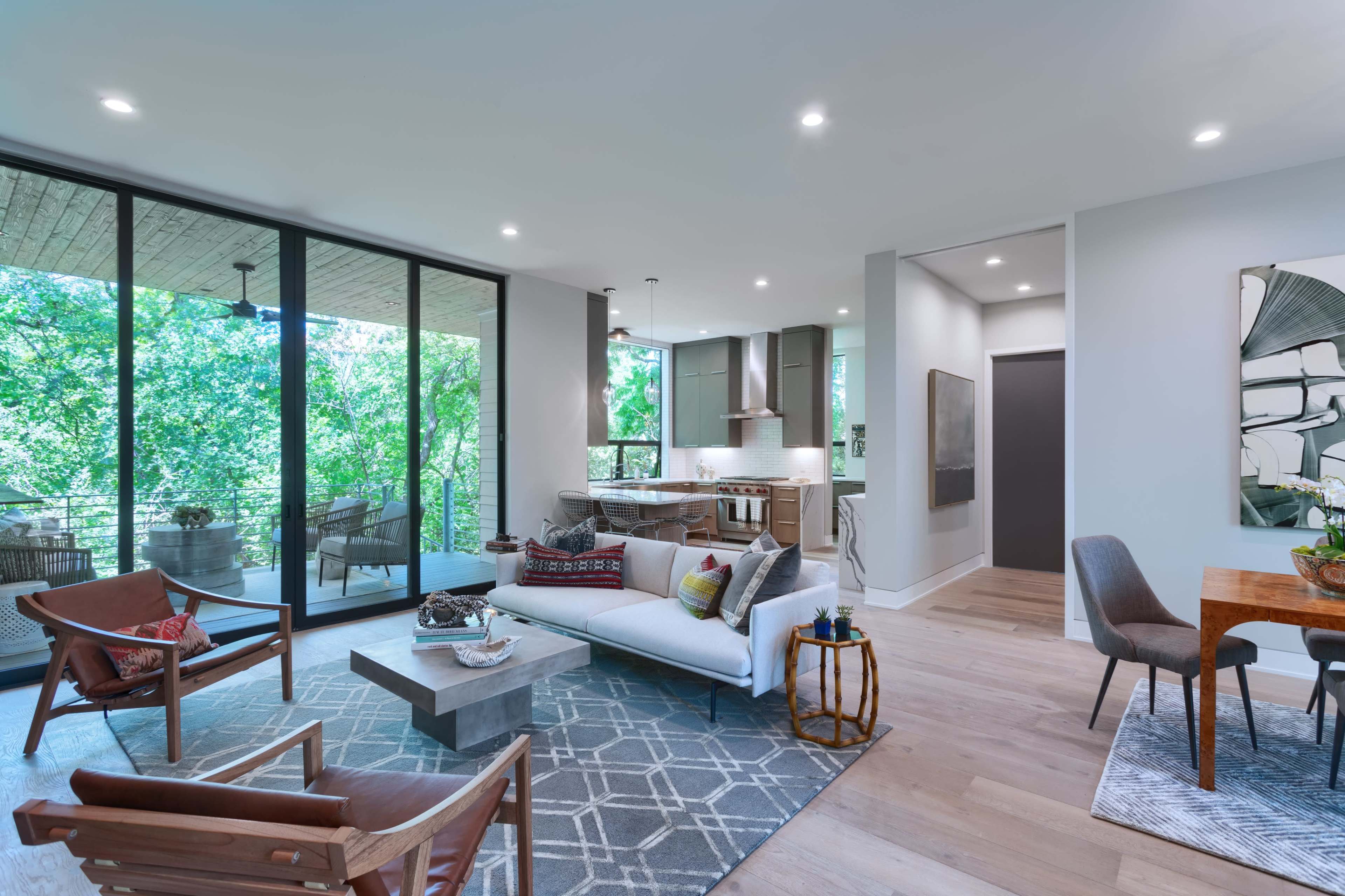 A modern living room with large windows overlooking greenery, featuring a seating area, a dining space, and a kitchen in the background.