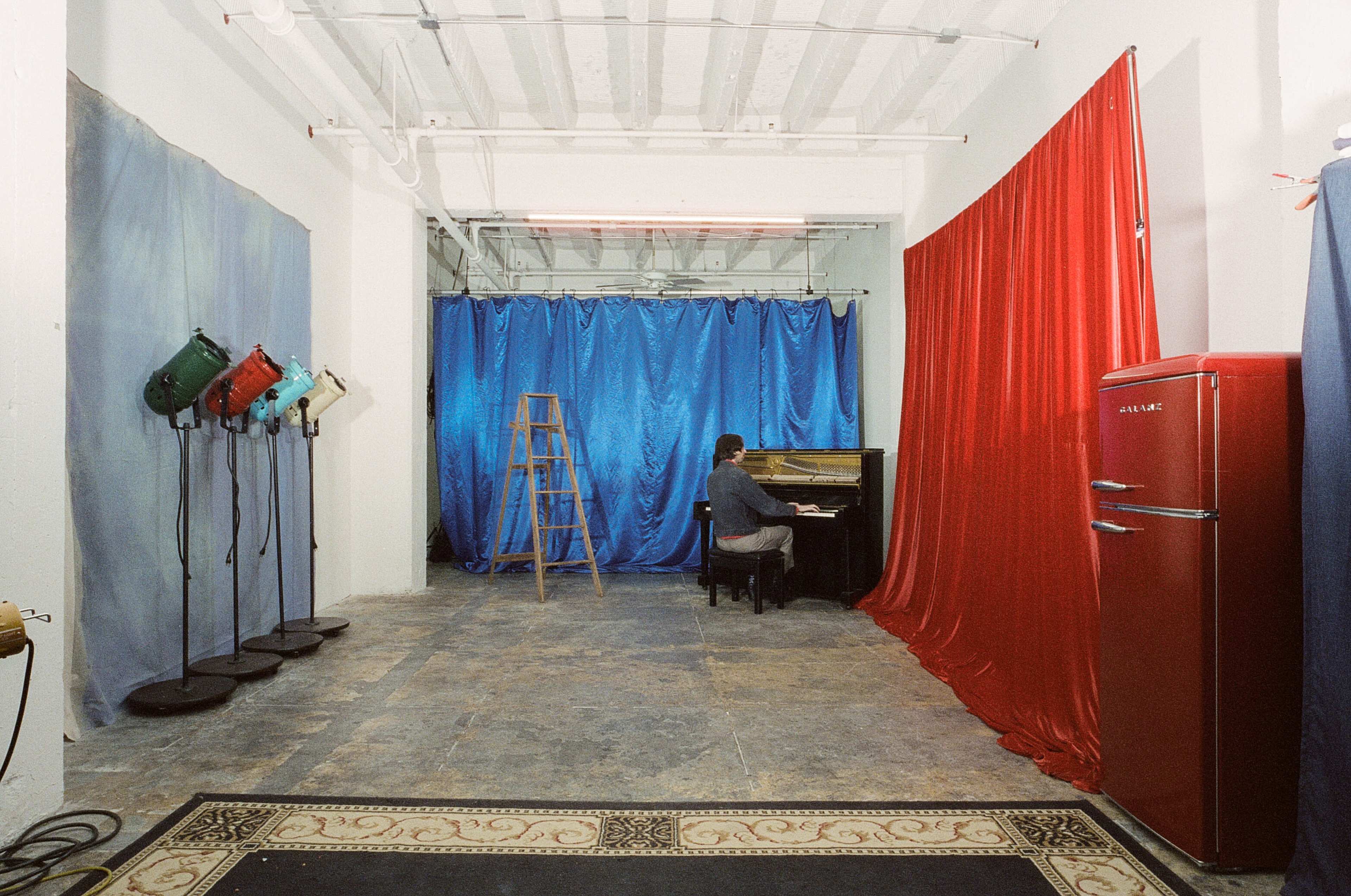 A person plays a piano in a room with blue and red curtains, vintage lighting equipment, a ladder, and a red refrigerator.