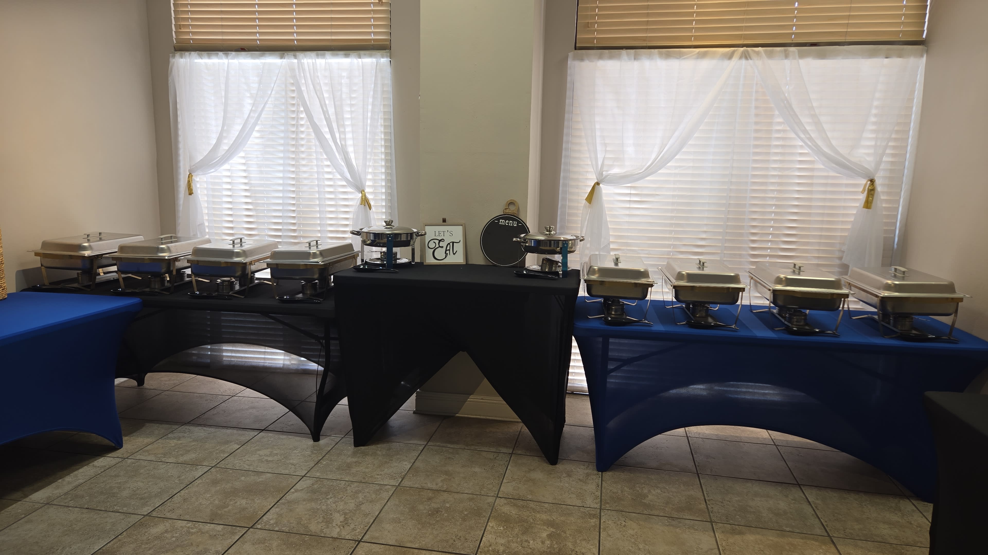 The image shows a catering setup with multiple chafing dishes arranged on black and blue tables, in front of large windows with white curtains.