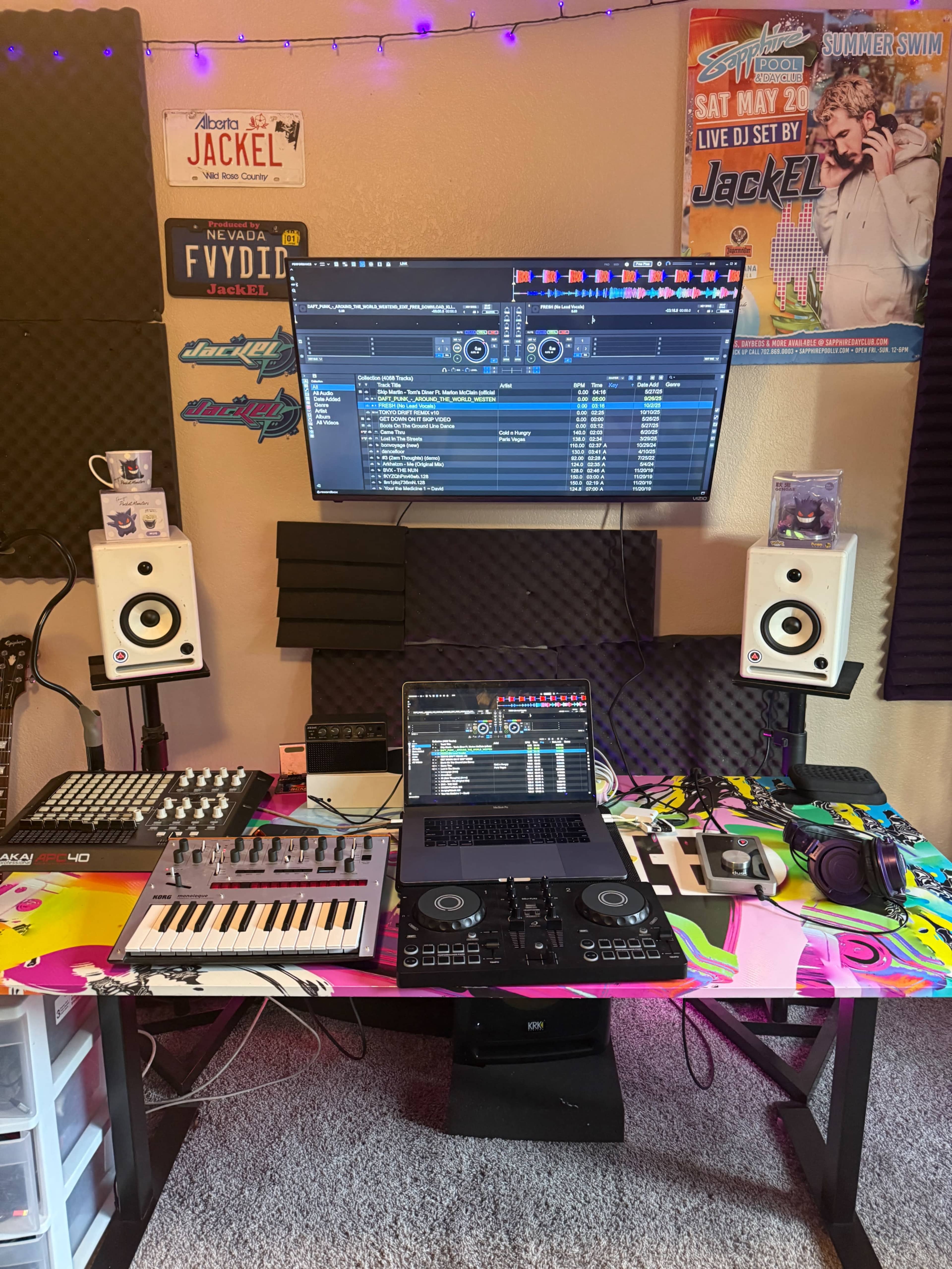 The image shows a music production setup featuring a laptop, a digital audio workstation on a large monitor, various music equipment, and speakers, all arranged on a colorful table.