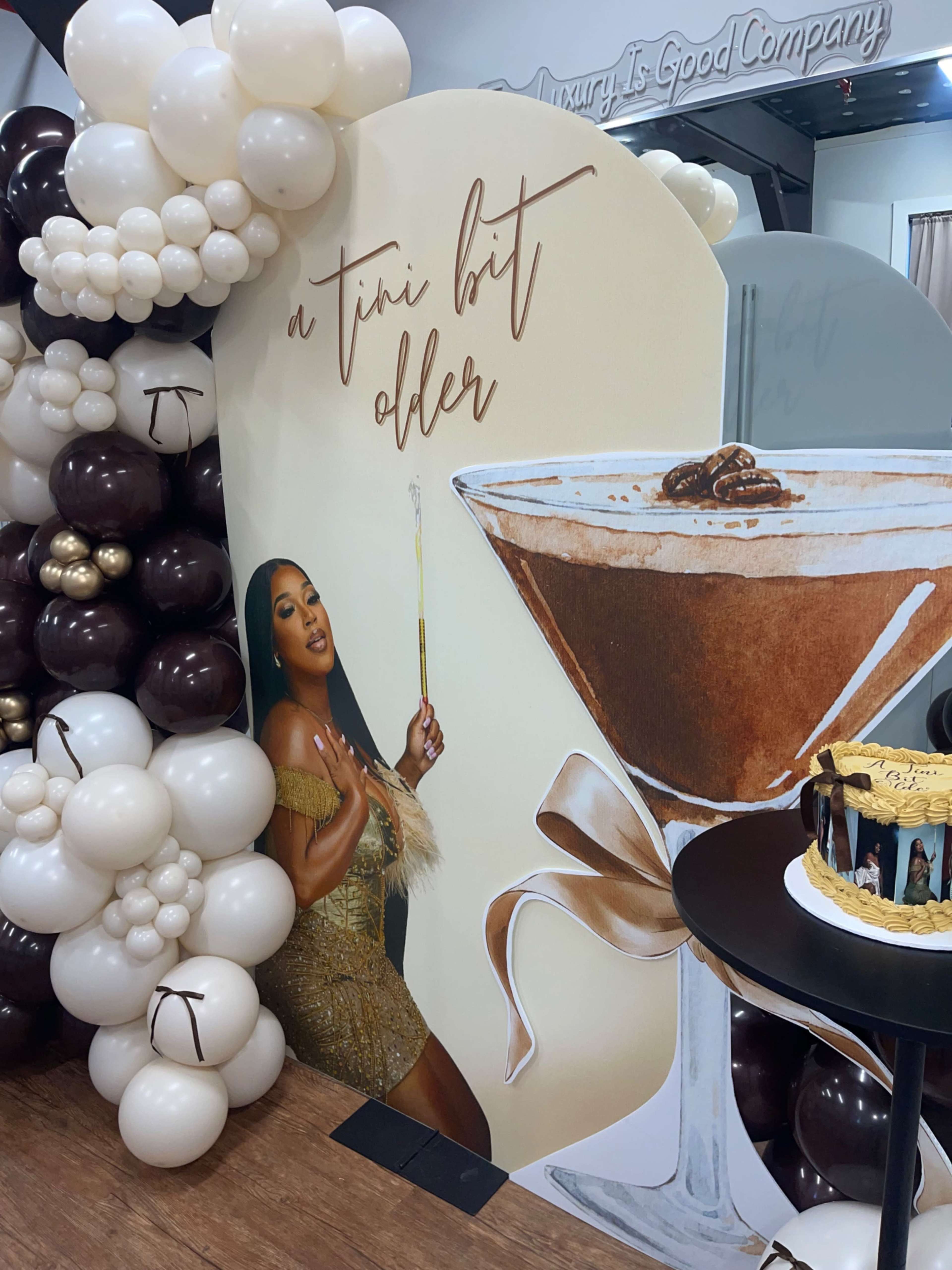 A large decorative display featuring a woman holding a cocktail is surrounded by balloons and a cake on a table.