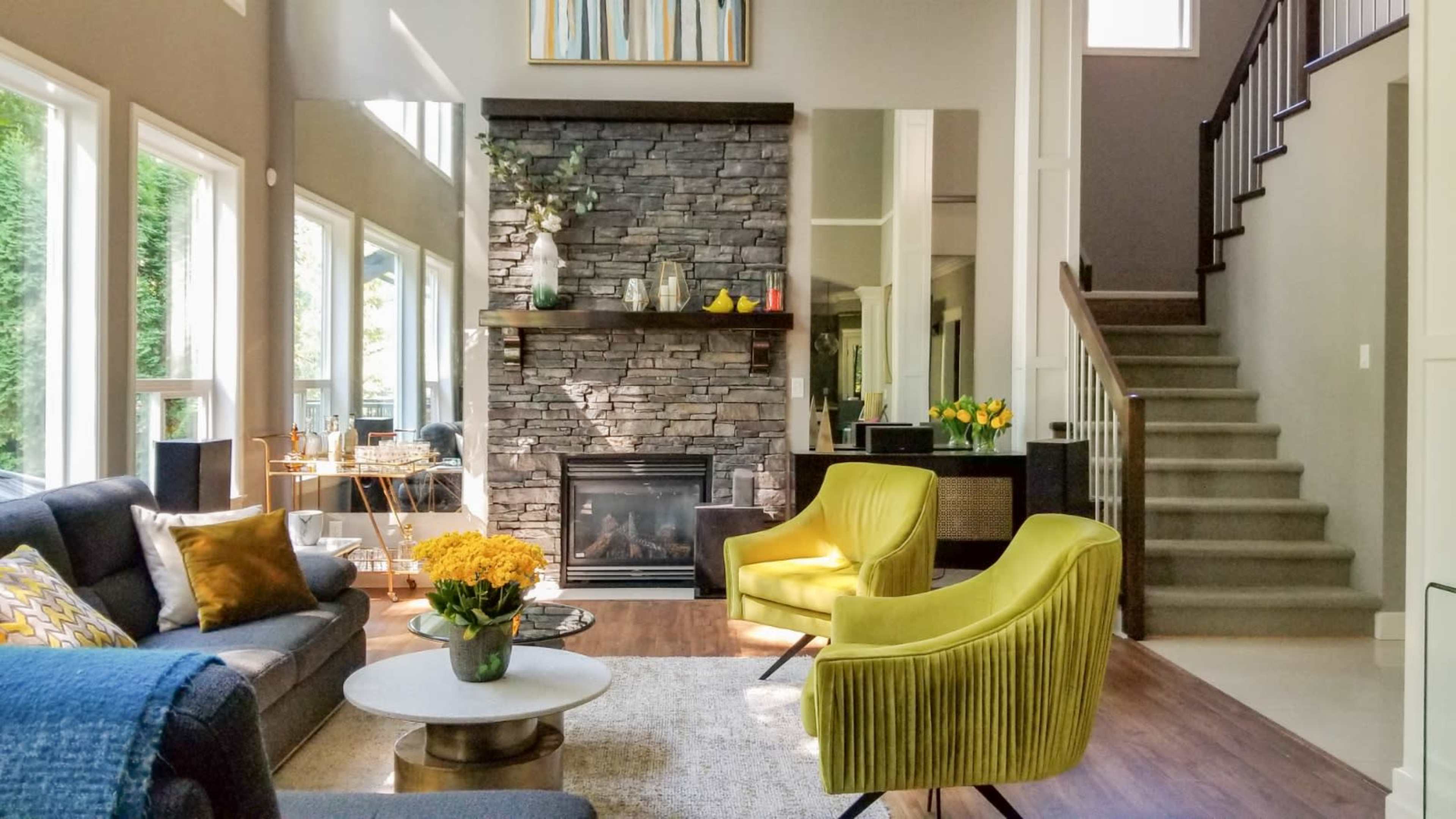 A modern living room with large windows, a stone fireplace, and yellow accent chairs, adjacent to a staircase.