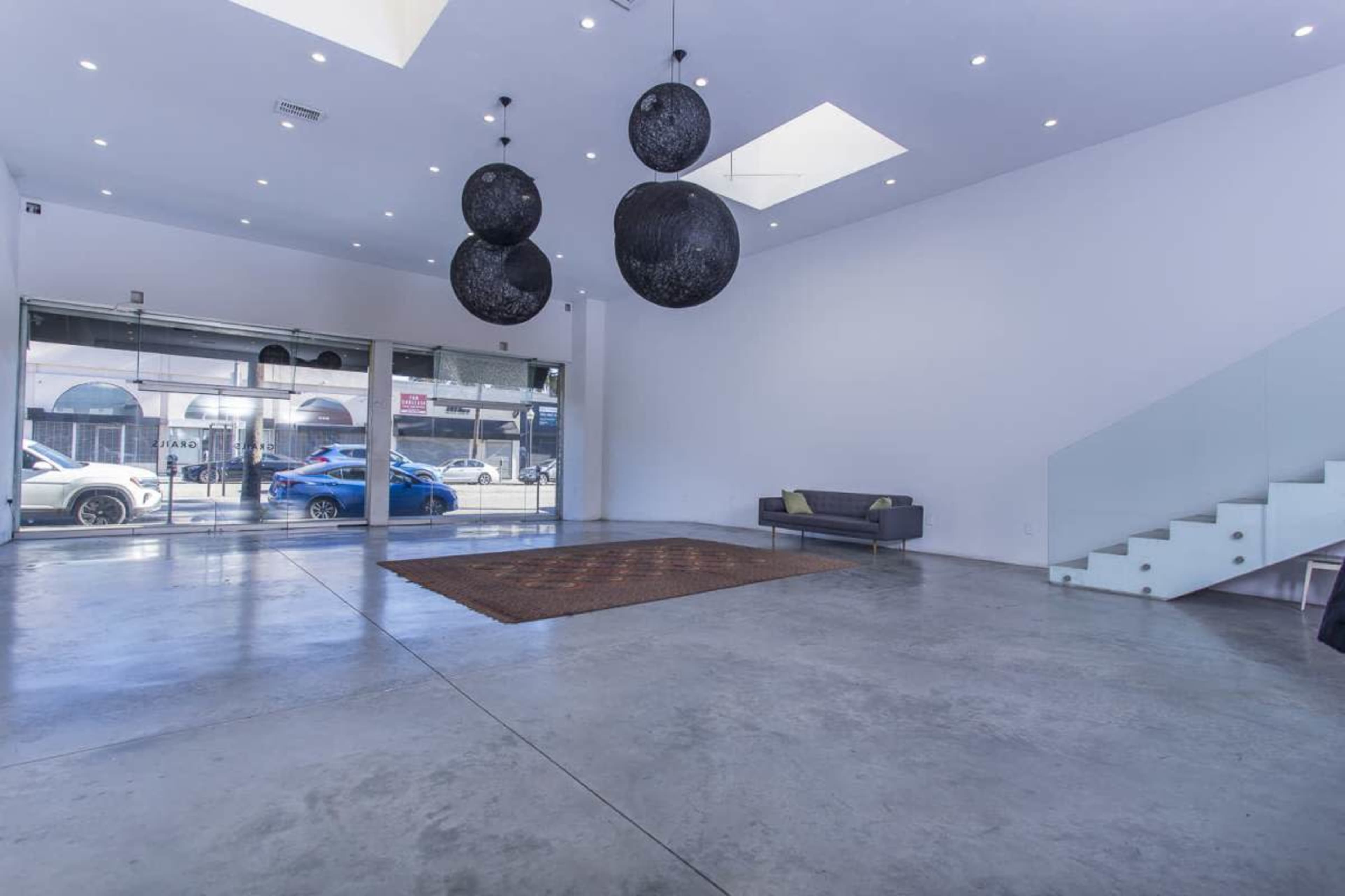 The image shows a spacious, modern interior with polished concrete floors, three large pendant lights hanging from the ceiling, a small sofa, and a staircase on one side.