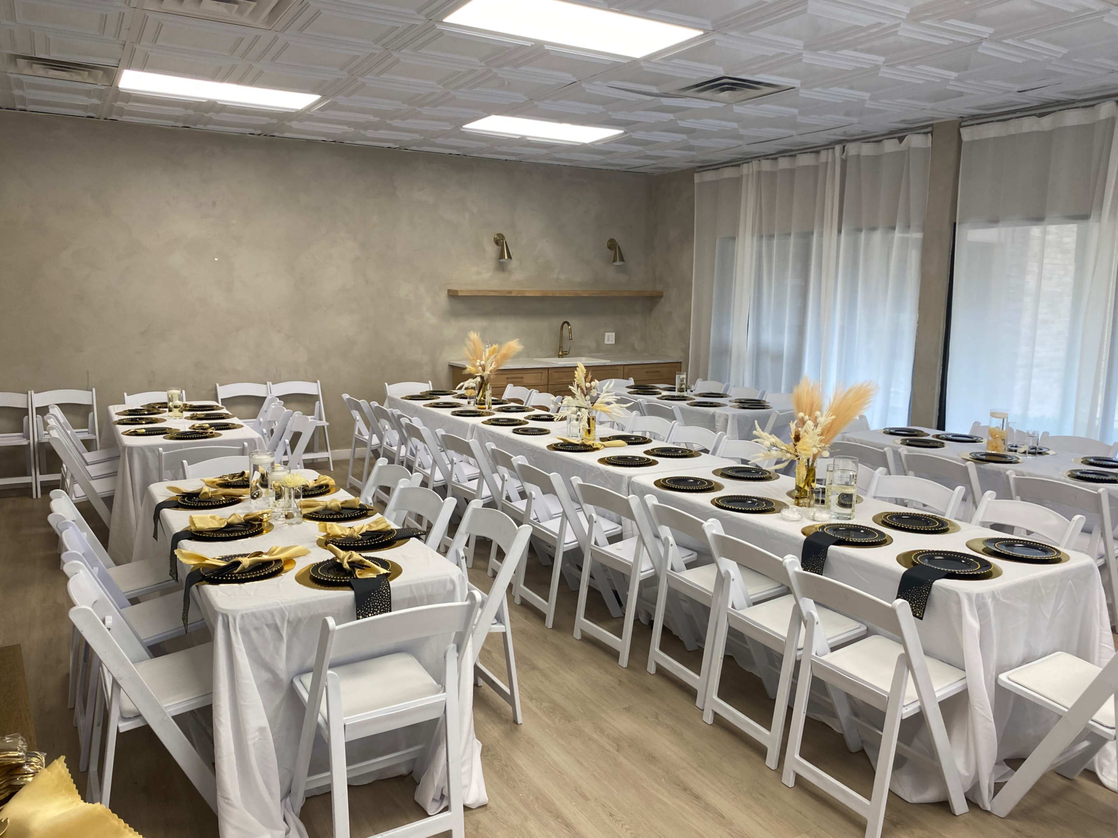 The image shows a well-lit banquet room set up with multiple rows of white tables and chairs, each adorned with black plates and gold cutlery, creating an elegant dining arrangement.