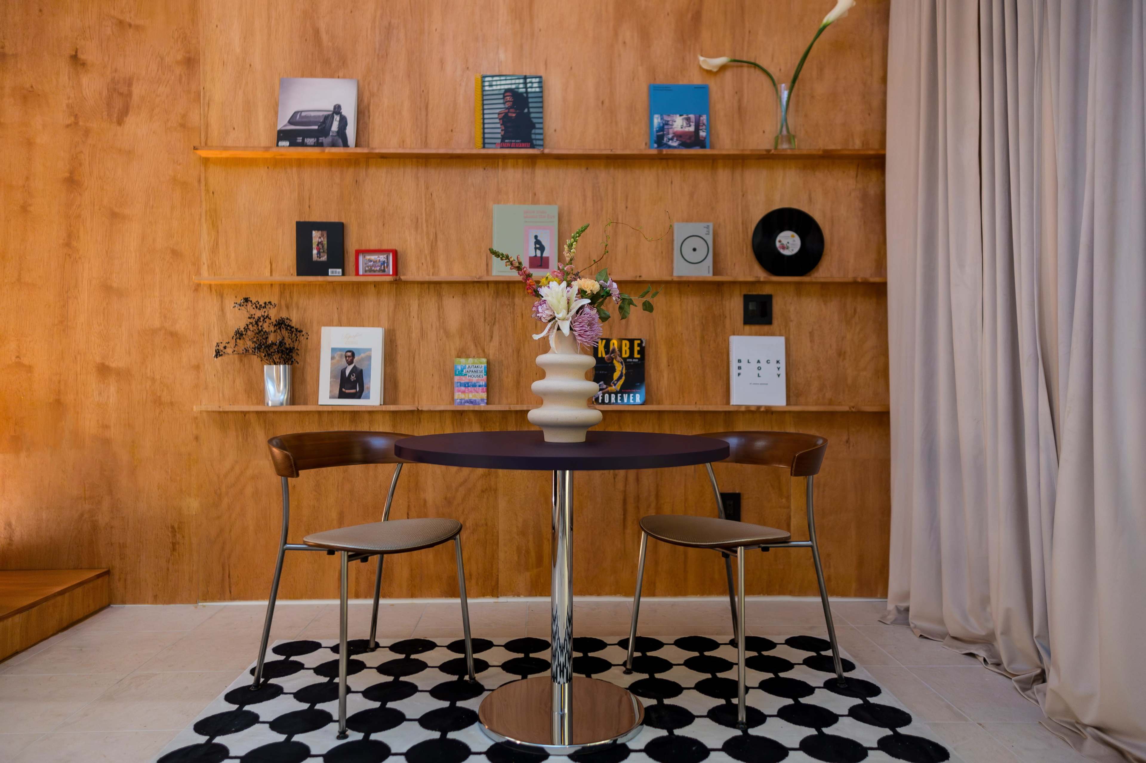 A round table with two chairs sits on a patterned rug in a room featuring a wooden wall with various framed items and a vase of flowers atop the table.