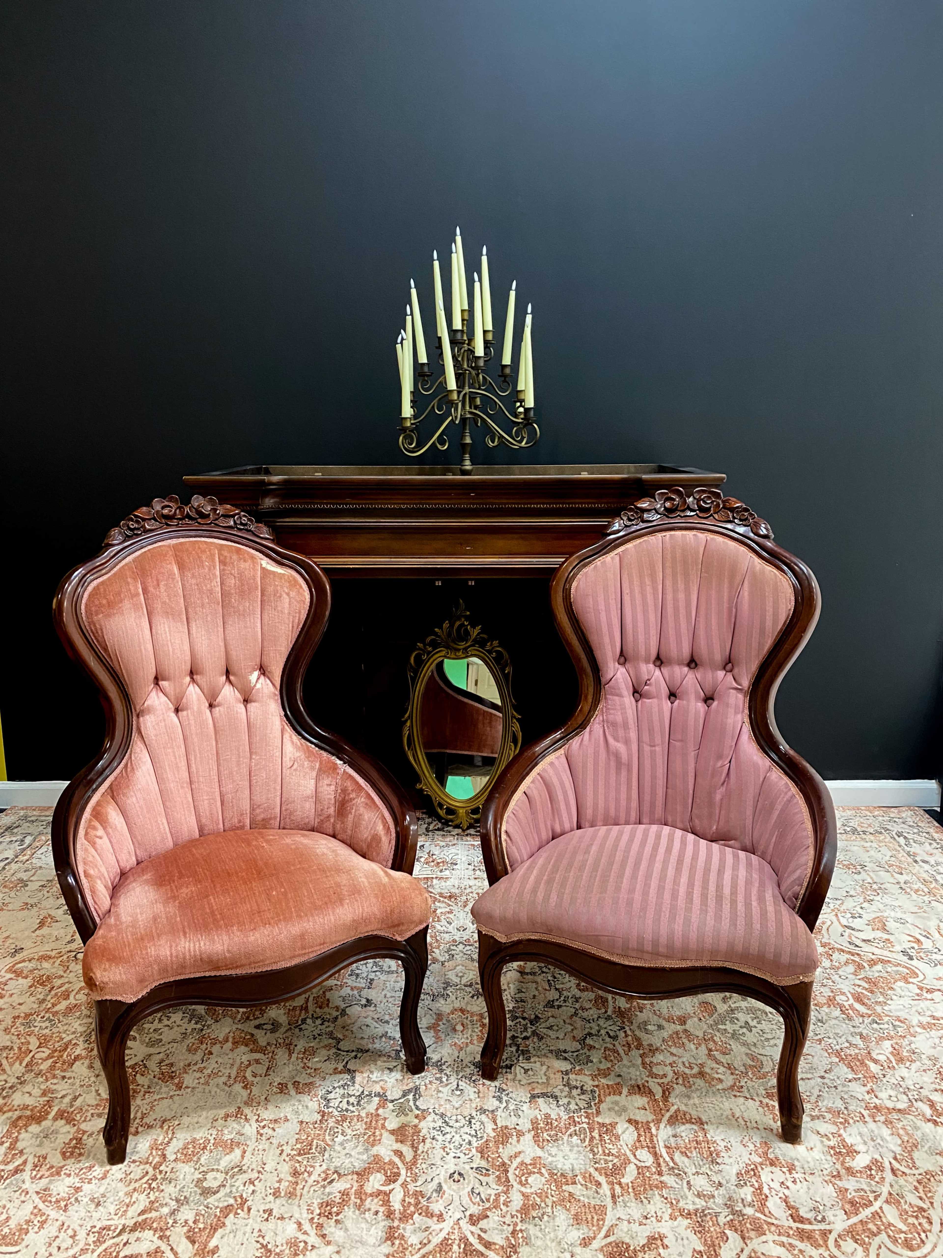 Two ornate upholstered chairs in pink tones are positioned facing each other in front of a dark wall, with a wooden console table and candelabra in the background.