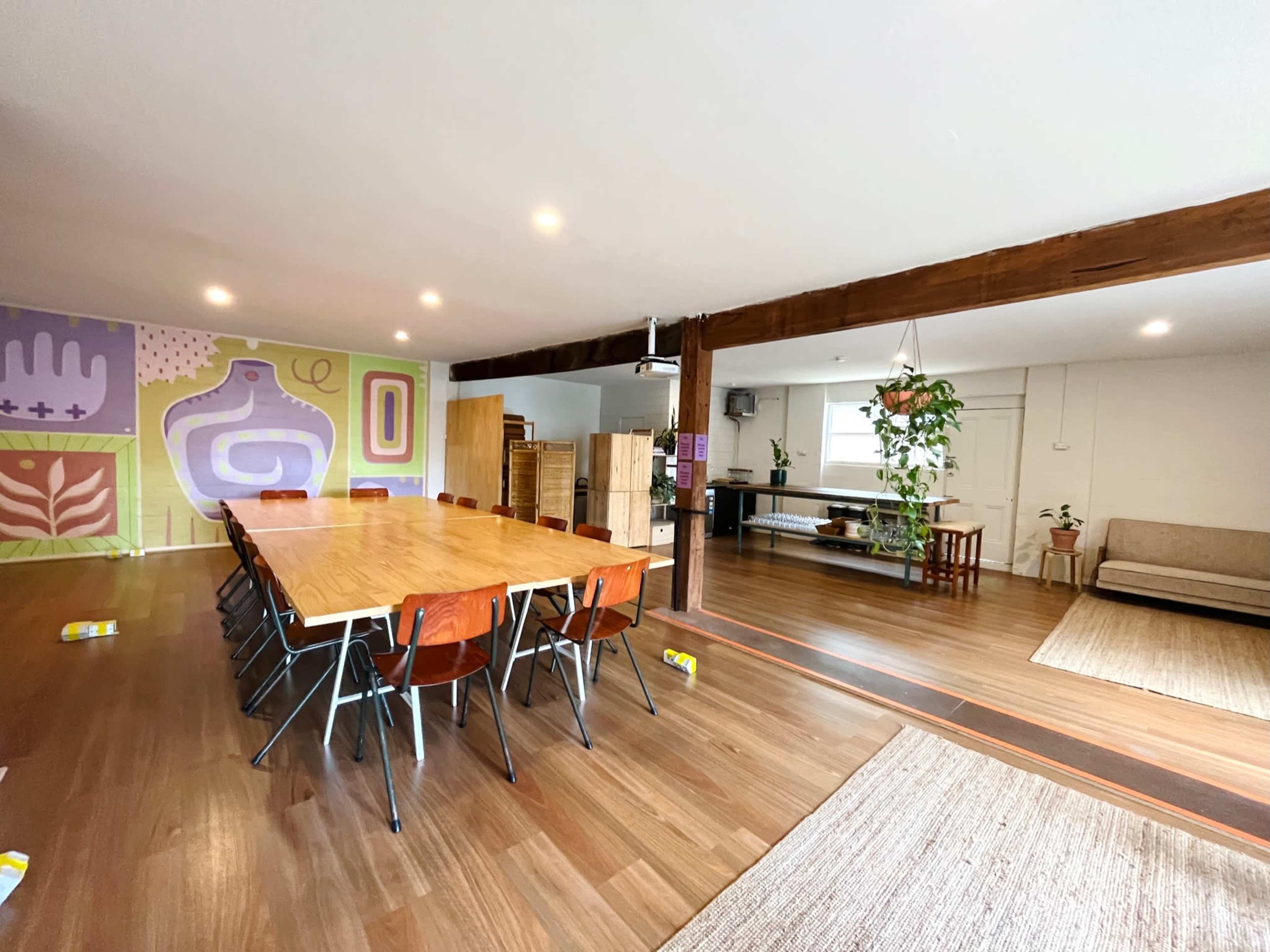 A spacious interior with a long wooden dining table surrounded by chairs on one side and a living area with plants and furniture on the other.