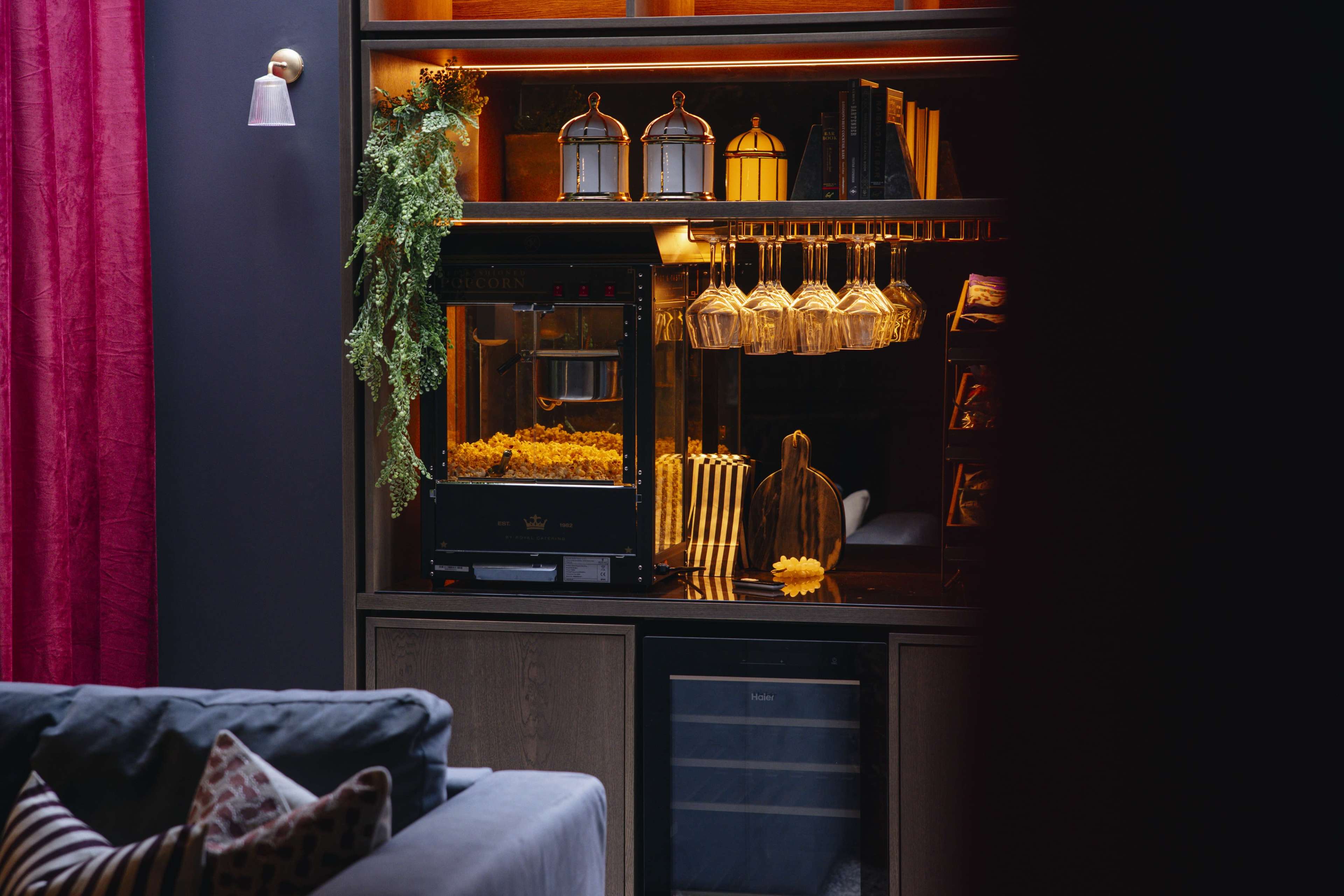 A modern entertainment area features a popcorn machine, glassware, and a small refrigerator set against a backdrop of dark walls and a plush sofa.