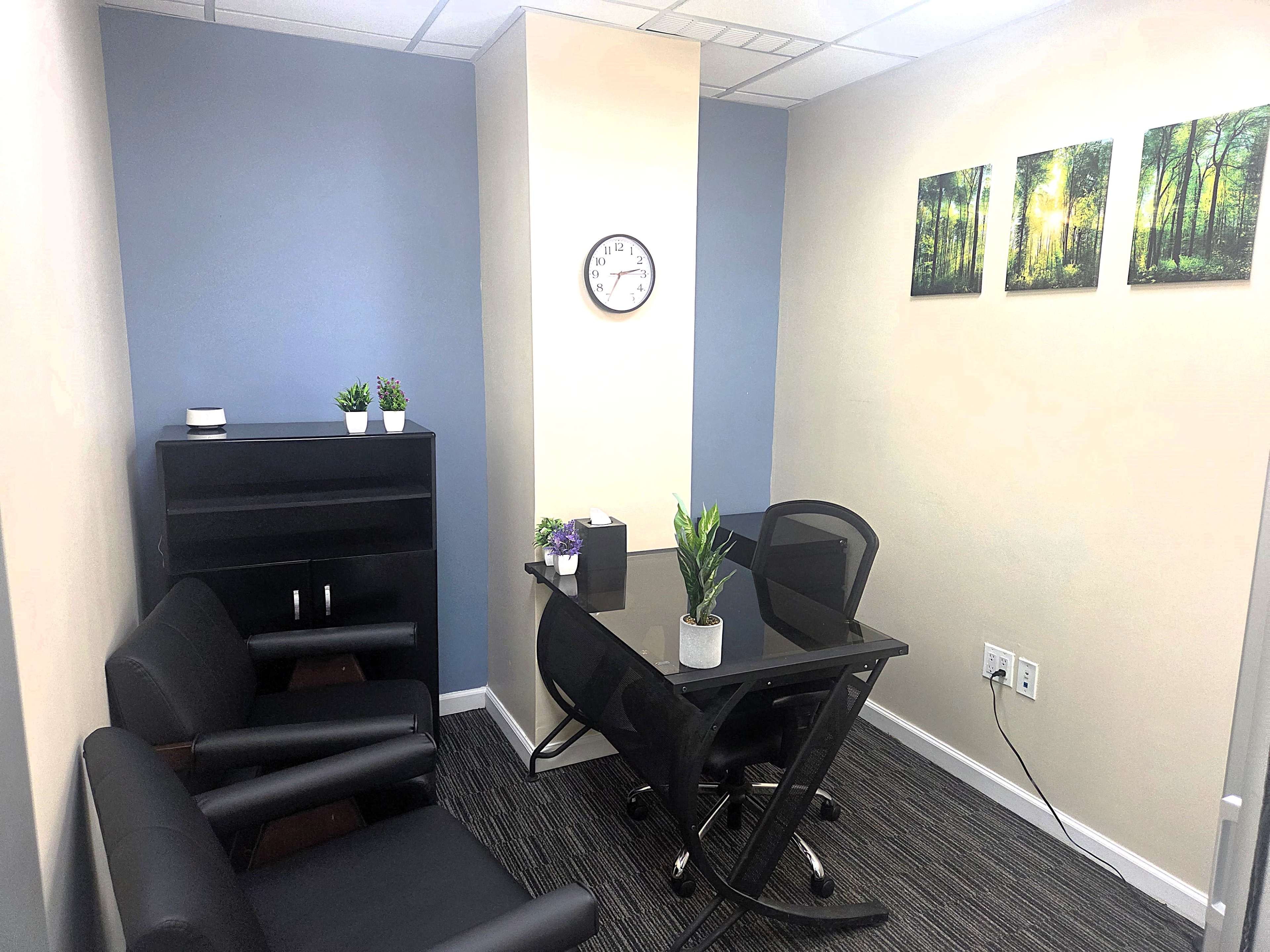 The image shows a small office space with a glass table, two black chairs, a clock on the wall, and framed pictures of trees.