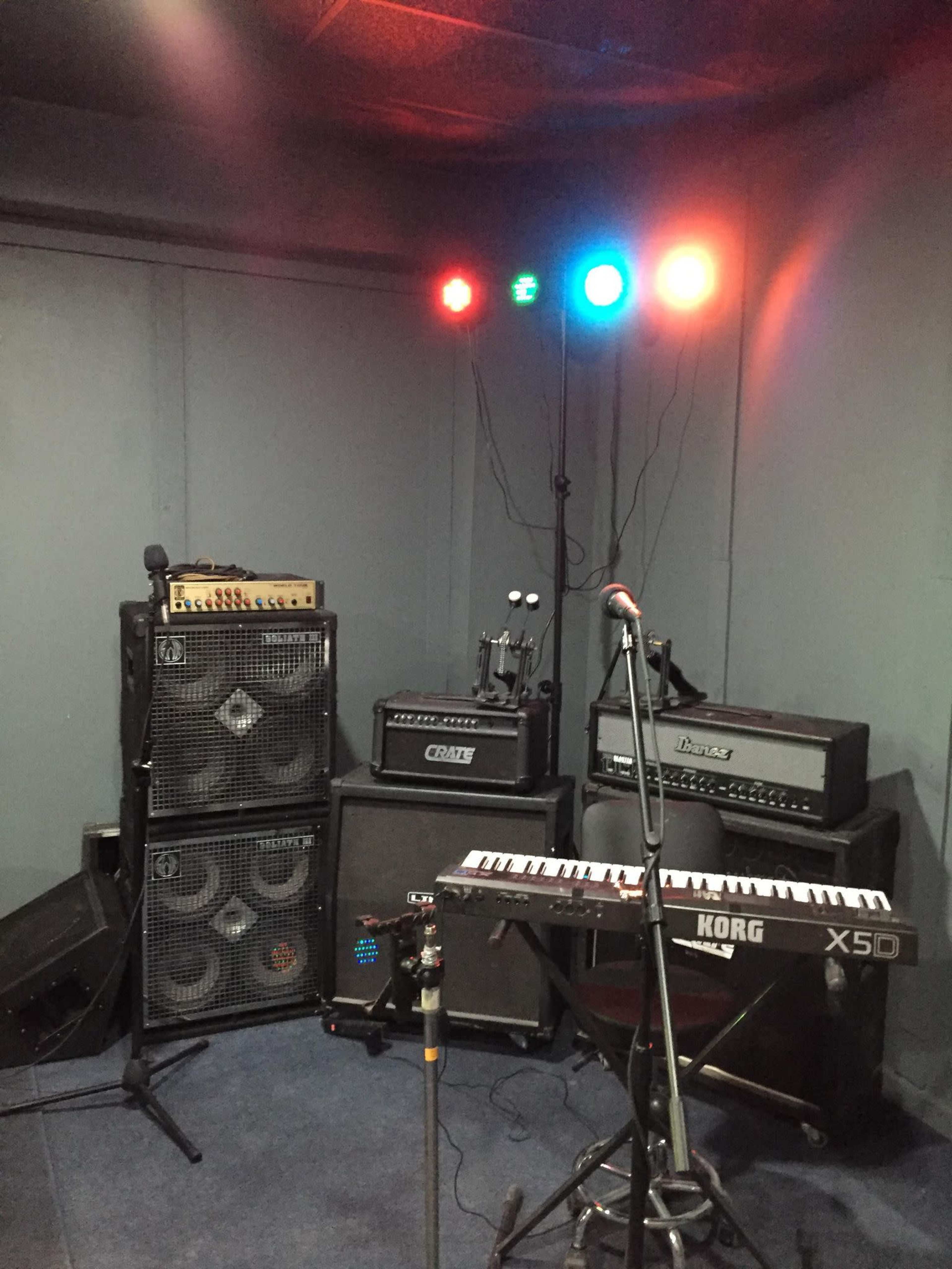 A small rehearsal room features several amplifiers and a keyboard set up under colored stage lights.