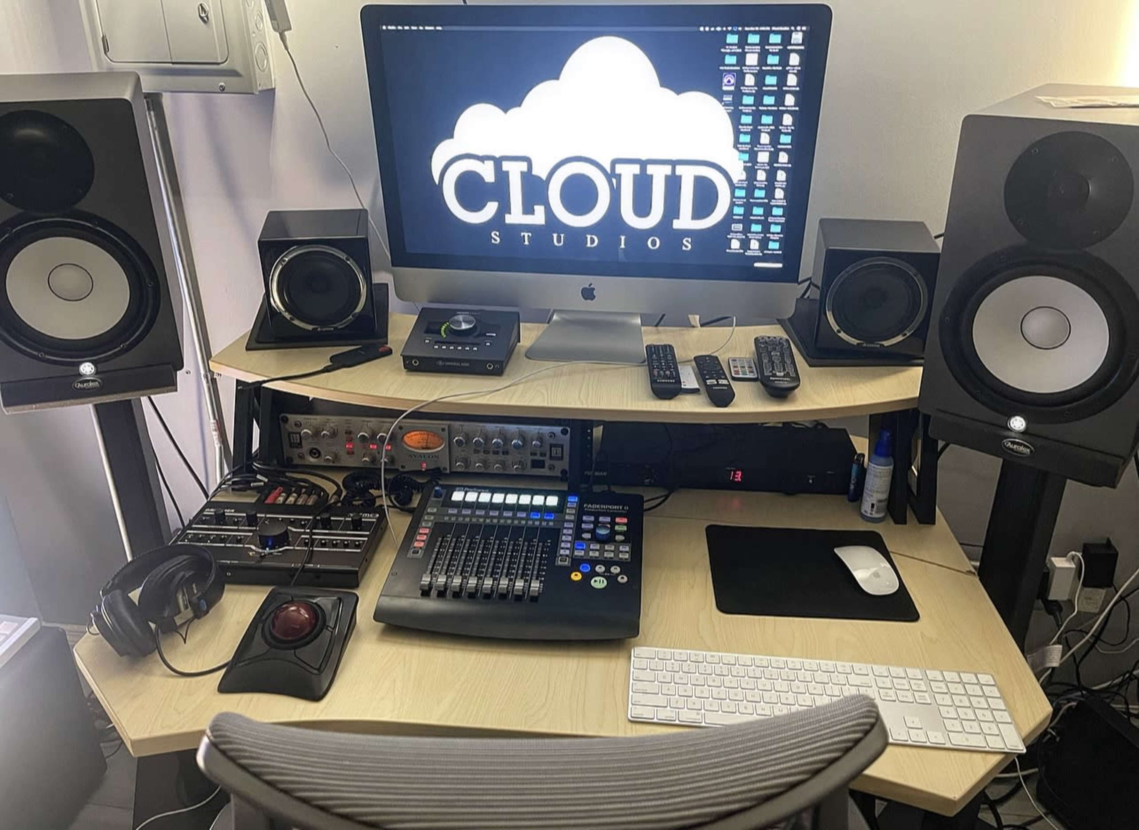 A home studio setup with a large computer monitor displaying "CLOUD STUDIOS," surrounded by audio equipment, speakers, mixers, and a keyboard.