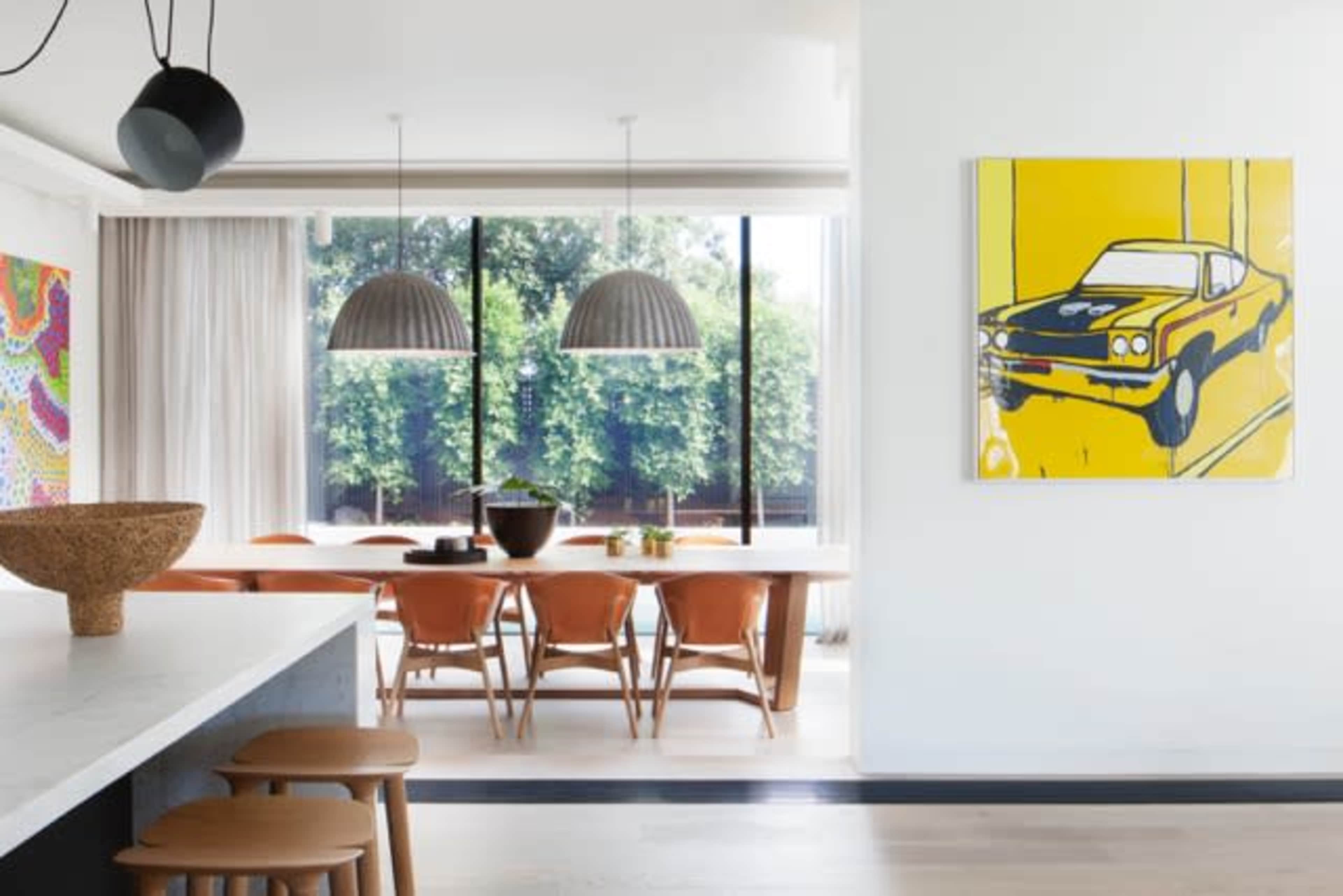 The image shows a modern dining area with a long wooden table surrounded by leather chairs, large windows letting in natural light, and a vibrant yellow artwork featuring a classic car on the wall.