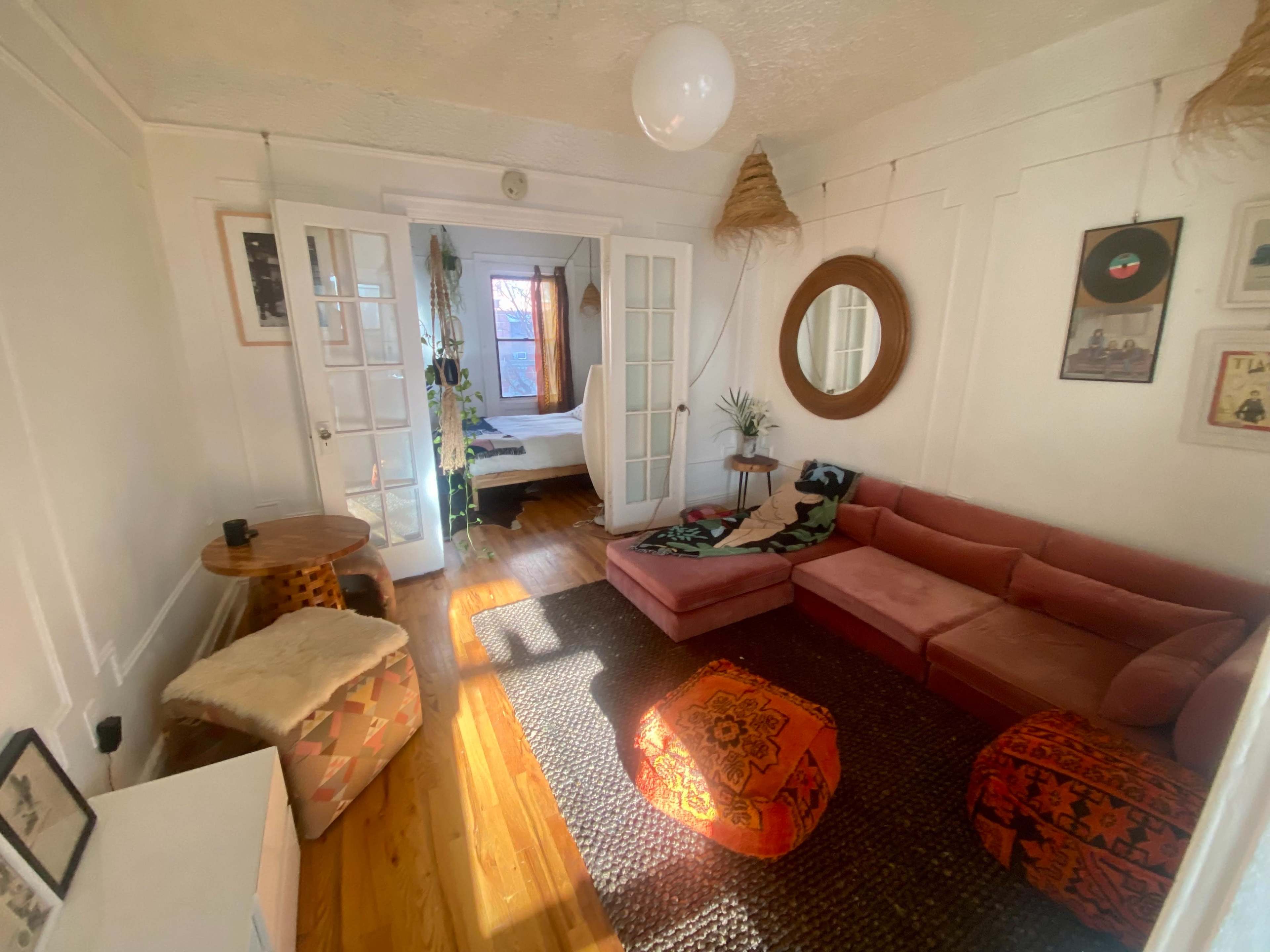 The image shows a cozy living room featuring a pink sofa, decorative poufs, a round mirror, and wooden flooring, with a view into a bedroom through glass doors.