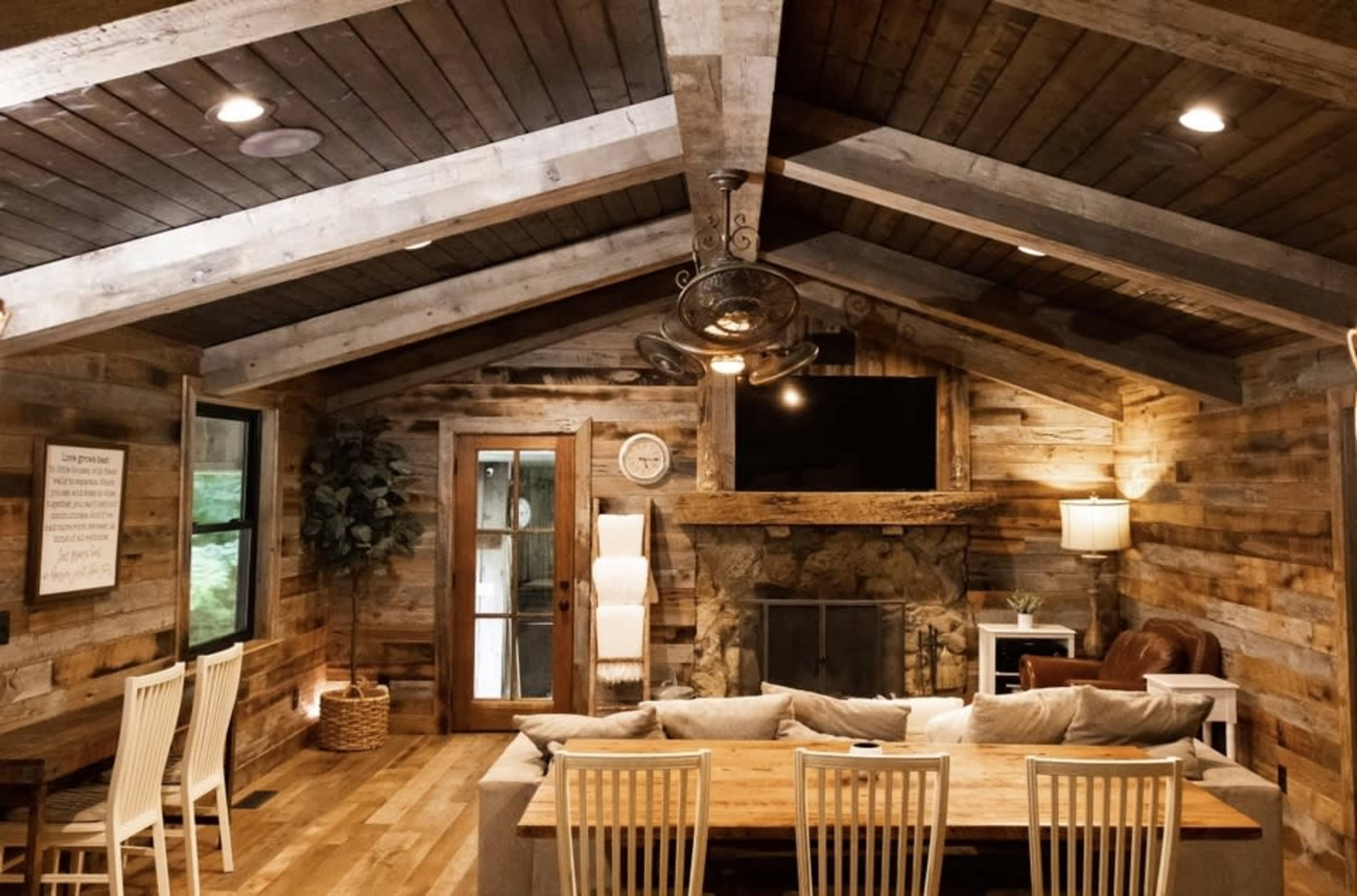 A rustic living room with wooden beams, a stone fireplace, and a large dining table surrounded by chairs.