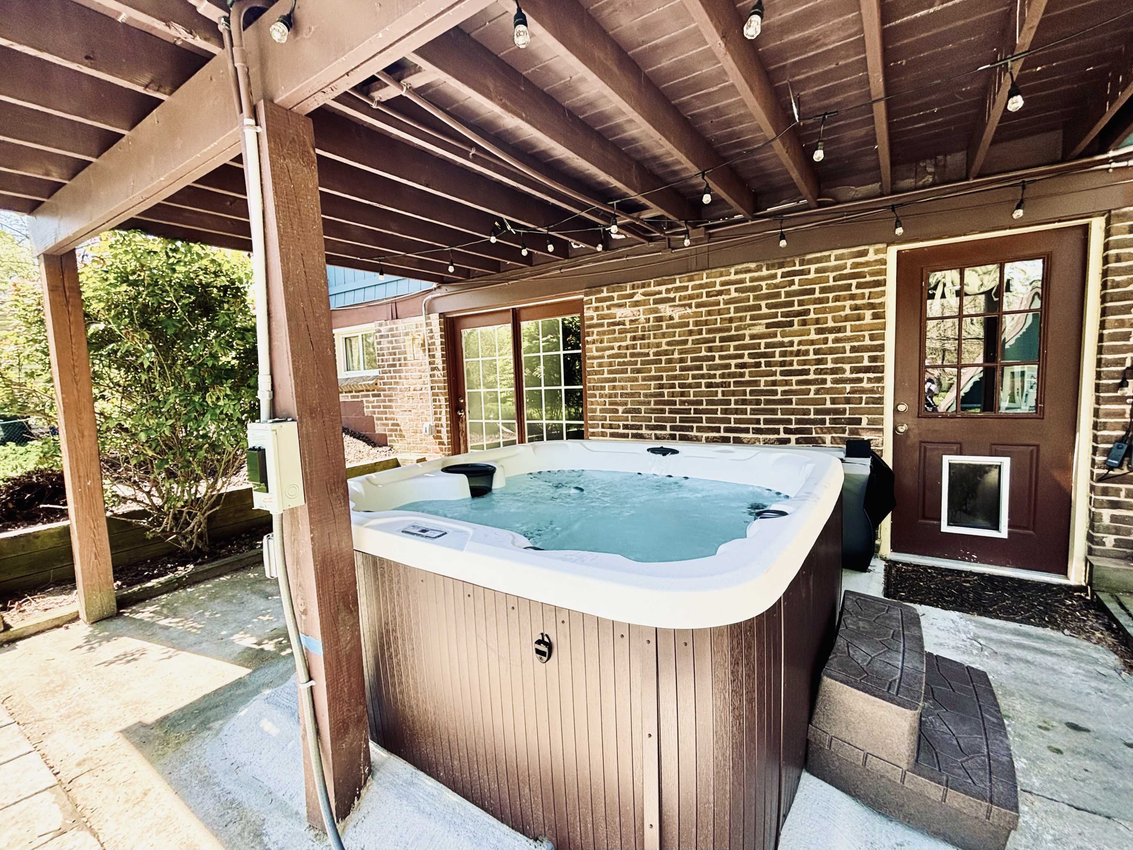 A hot tub located under a covered patio next to a brick wall and a door leading to a house.