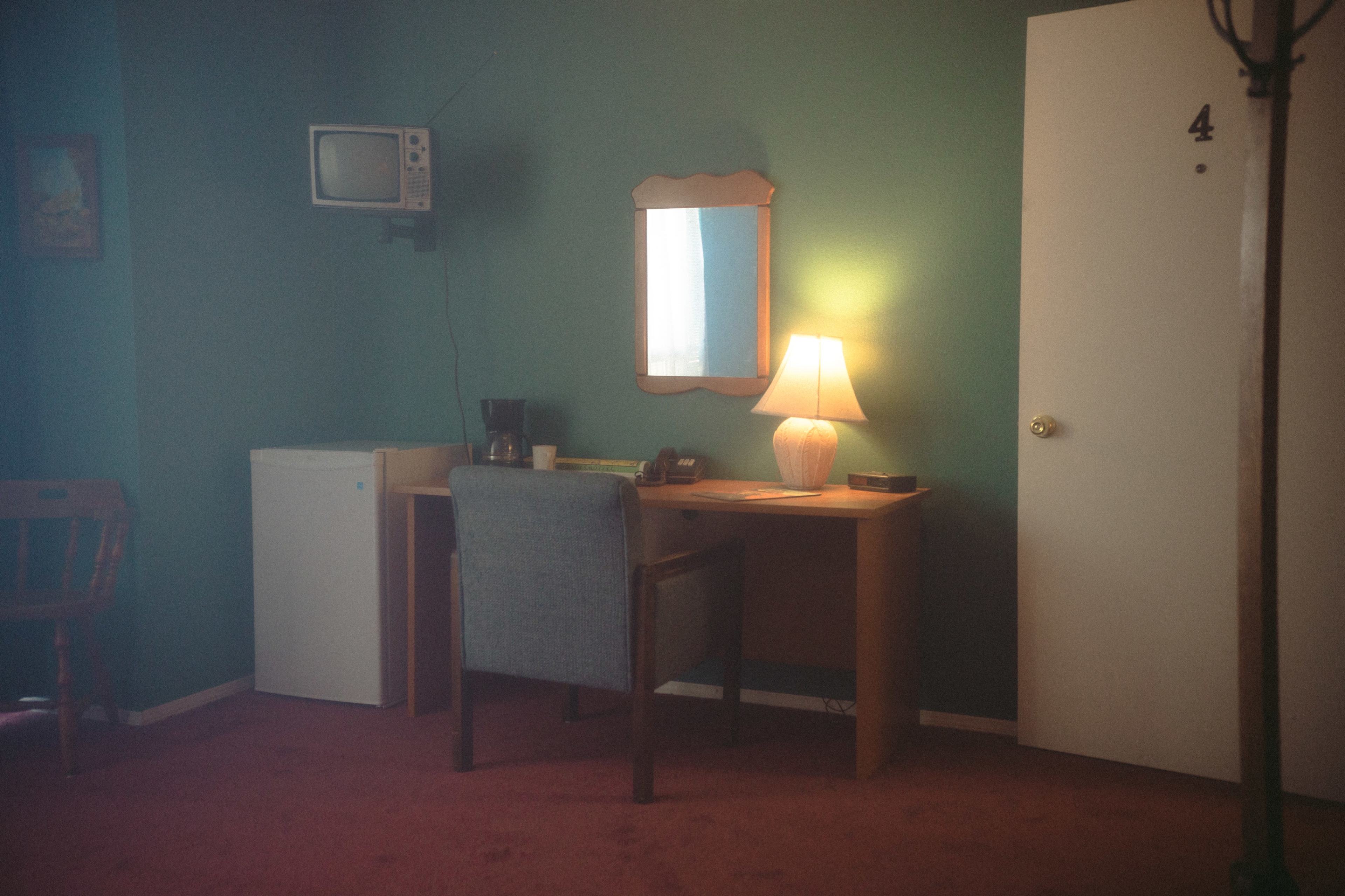 The image shows a small, simple hotel room with a green wall, a desk with a chair, a lamp, a mirror, a television, a refrigerator, and a door numbered four.