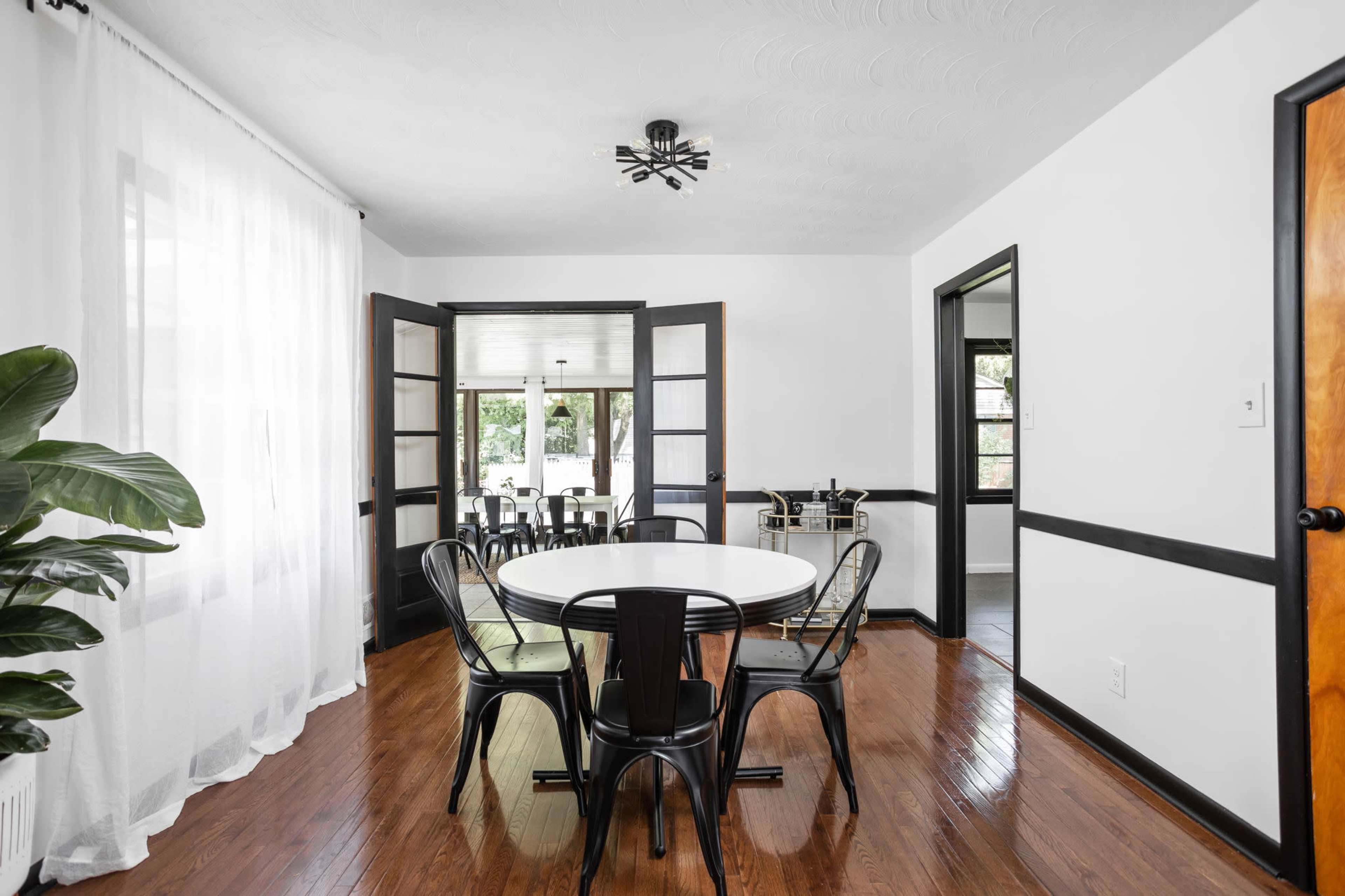 A dining room features a round table surrounded by black chairs, with French doors leading to an adjacent space illuminated by large windows.