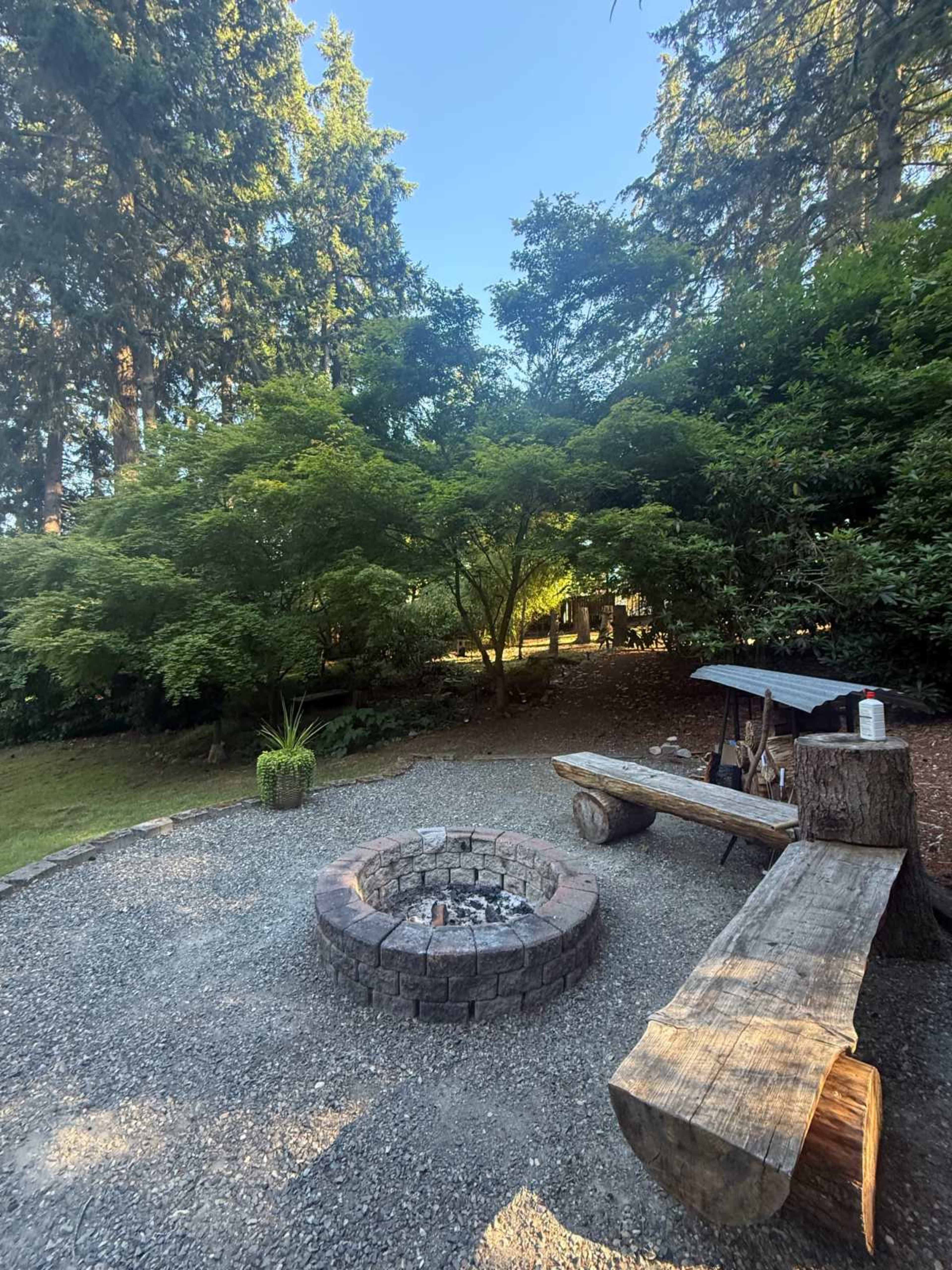 The image shows a fire pit made of stone surrounded by gravel, with a wooden bench nearby and lush greenery from trees and shrubs in the background.