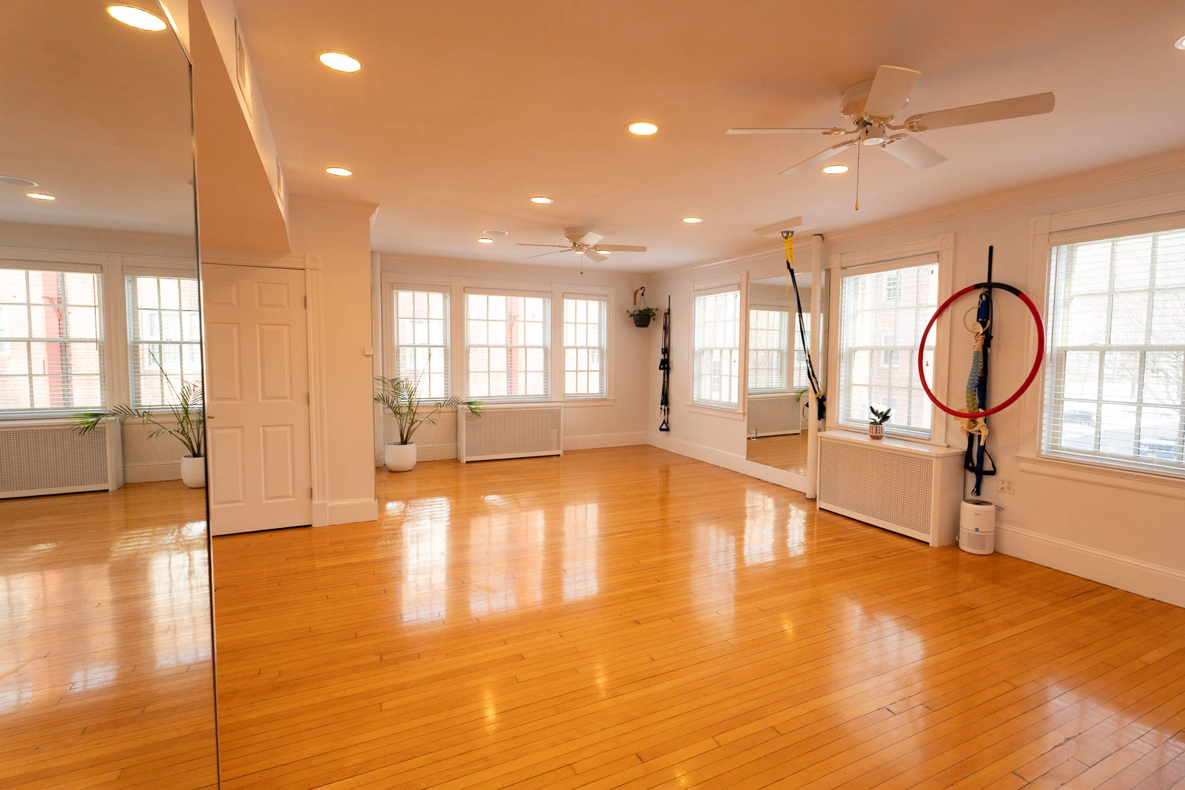 The image shows a spacious room with wooden flooring, large windows, and mirrors along one wall.