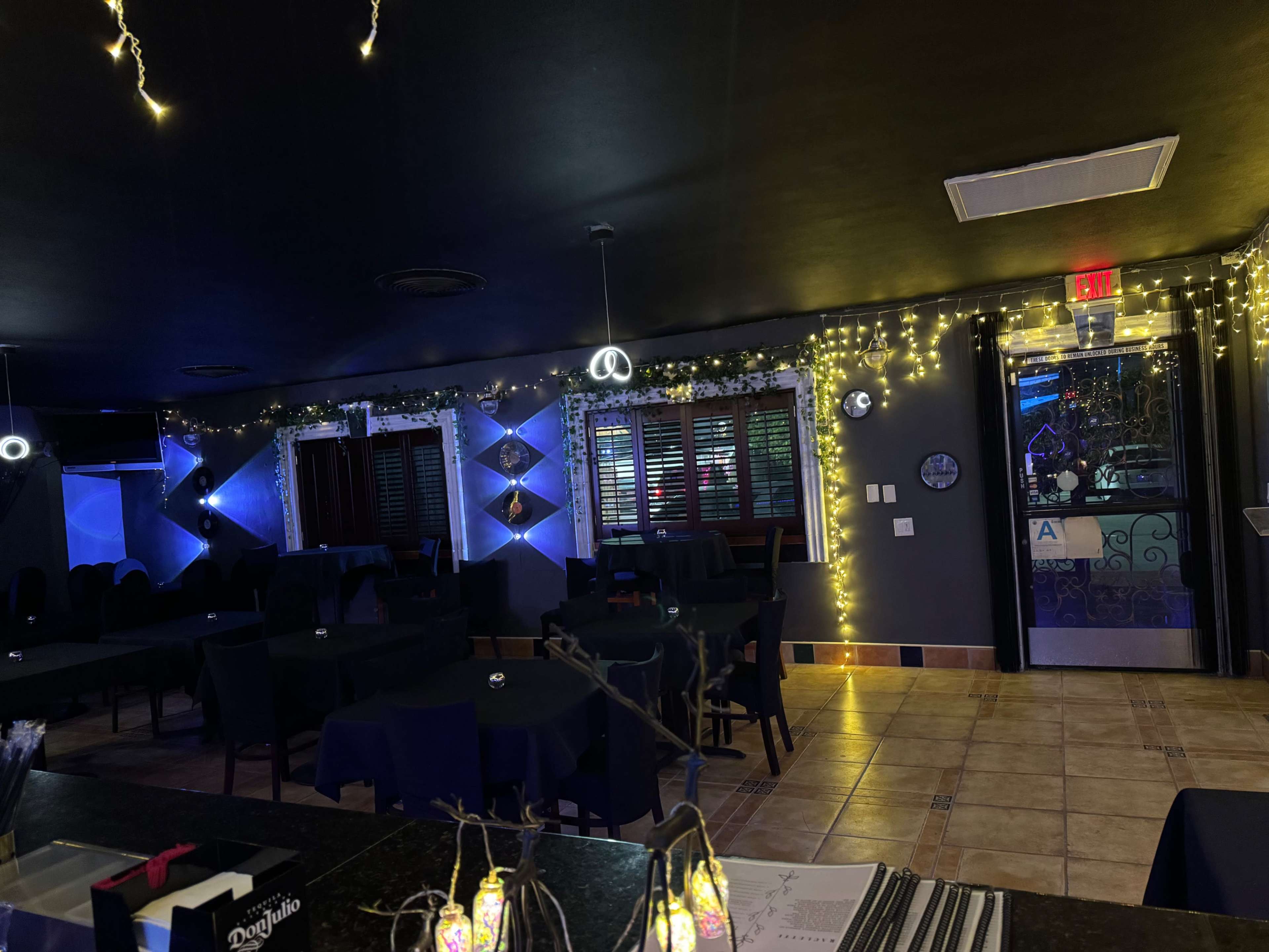 An interior of a dimly lit restaurant with dark tables, string lights along the walls, and a door leading outside.