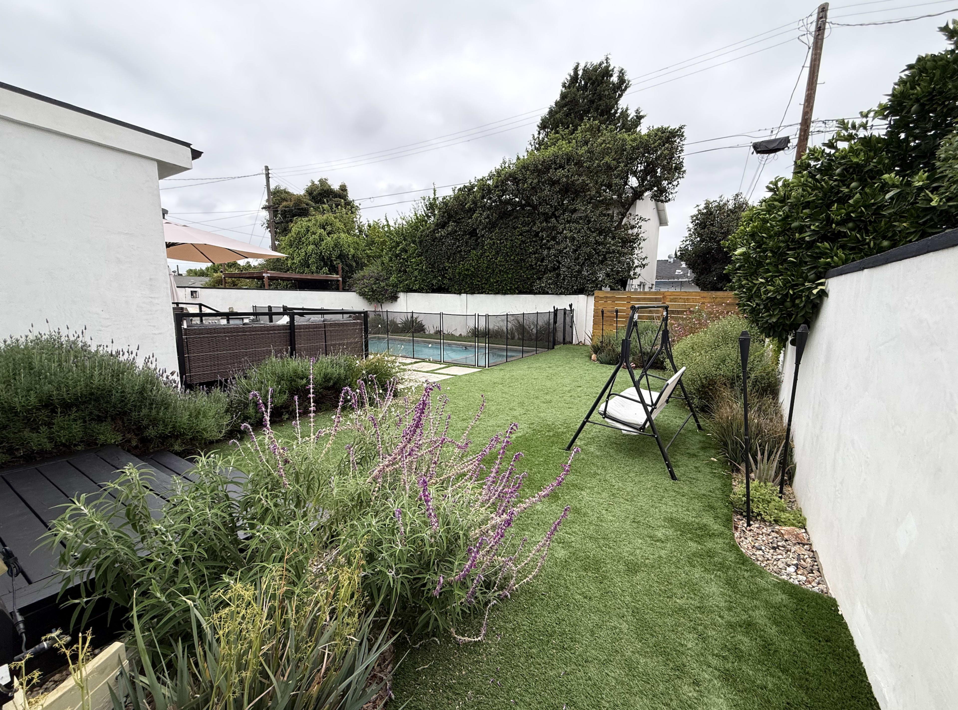 The image shows a landscaped backyard with a lawn, a swing seat, and a fenced pool area surrounded by greenery and decorative stones.