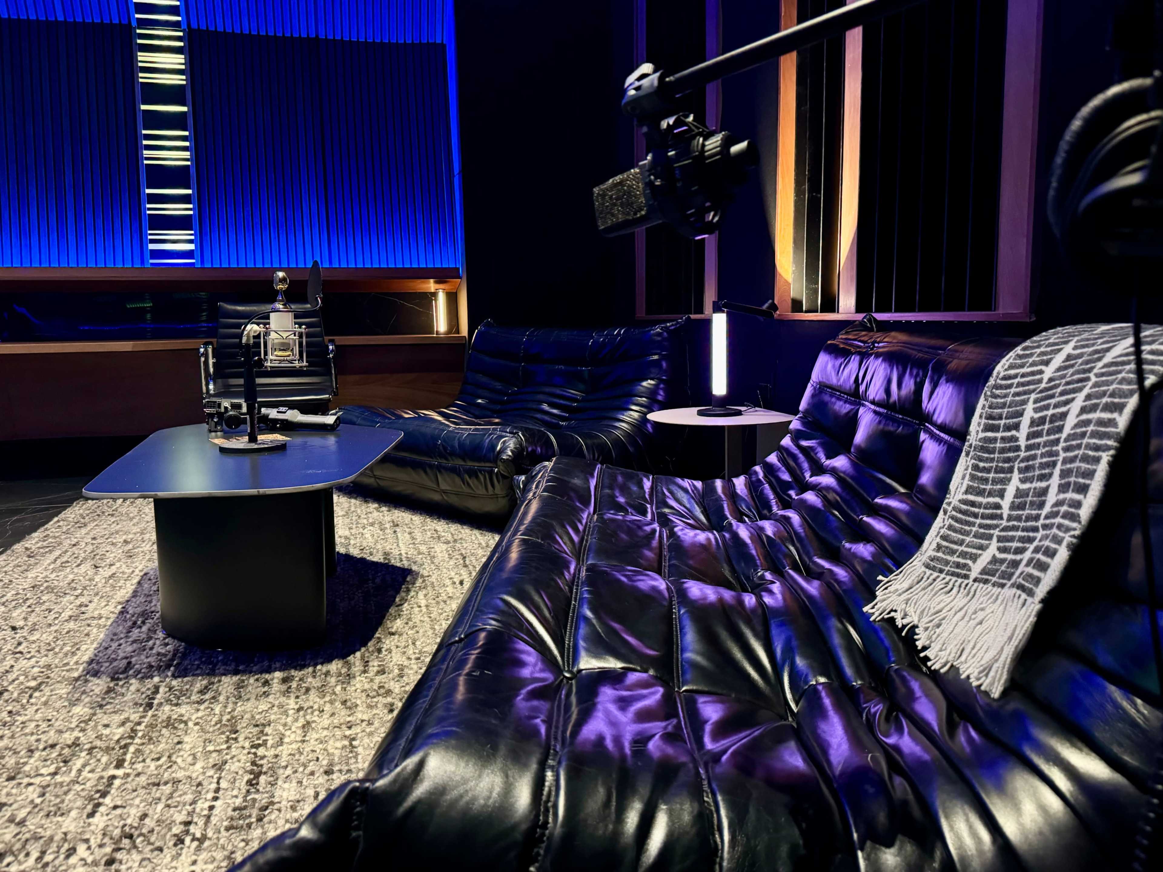A modern, minimalist lounge area featuring two black, tufted sofas and a small table, set against a backdrop of blue lighting and sound panels.