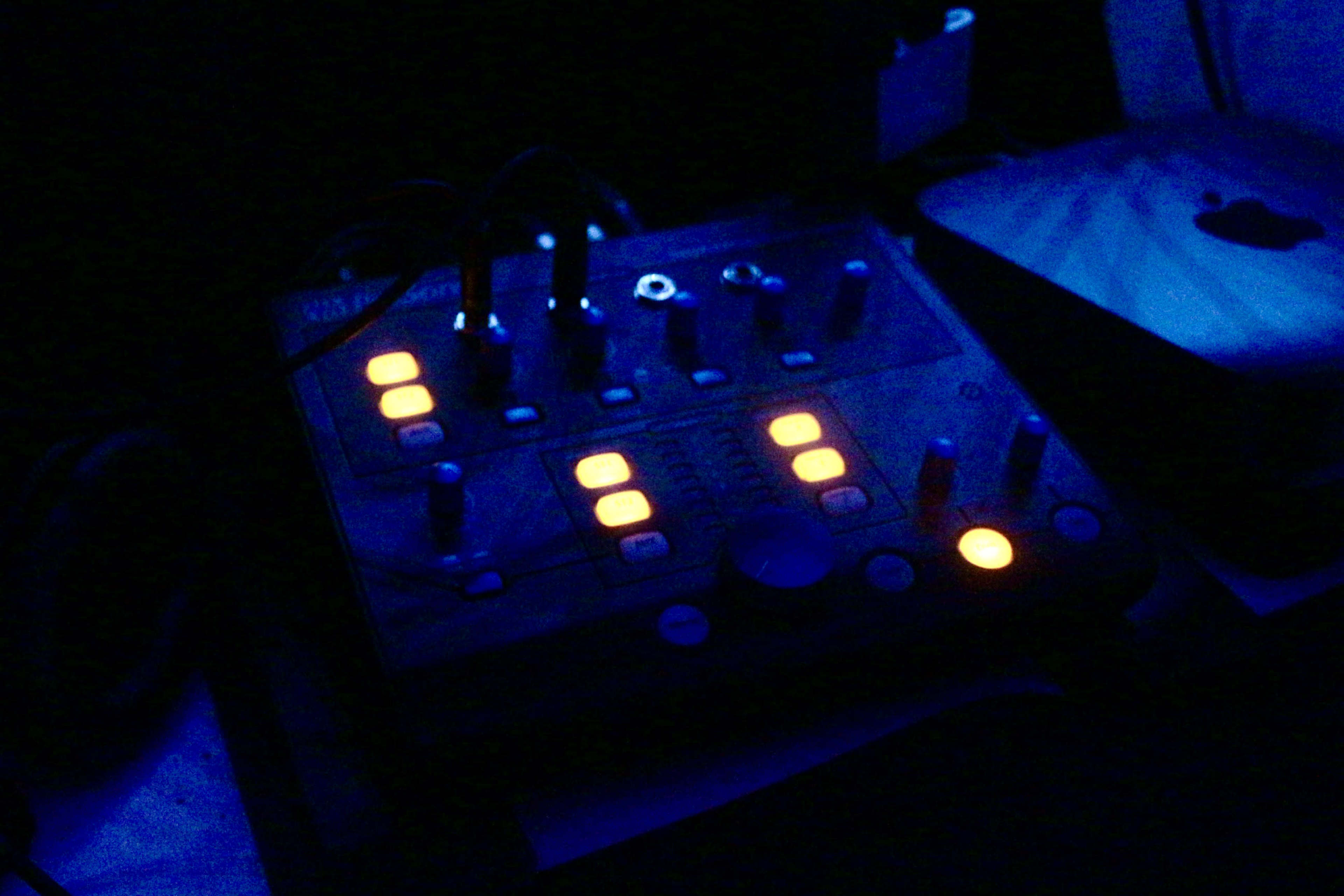 A dark room features a music mixer with illuminated buttons and knobs.