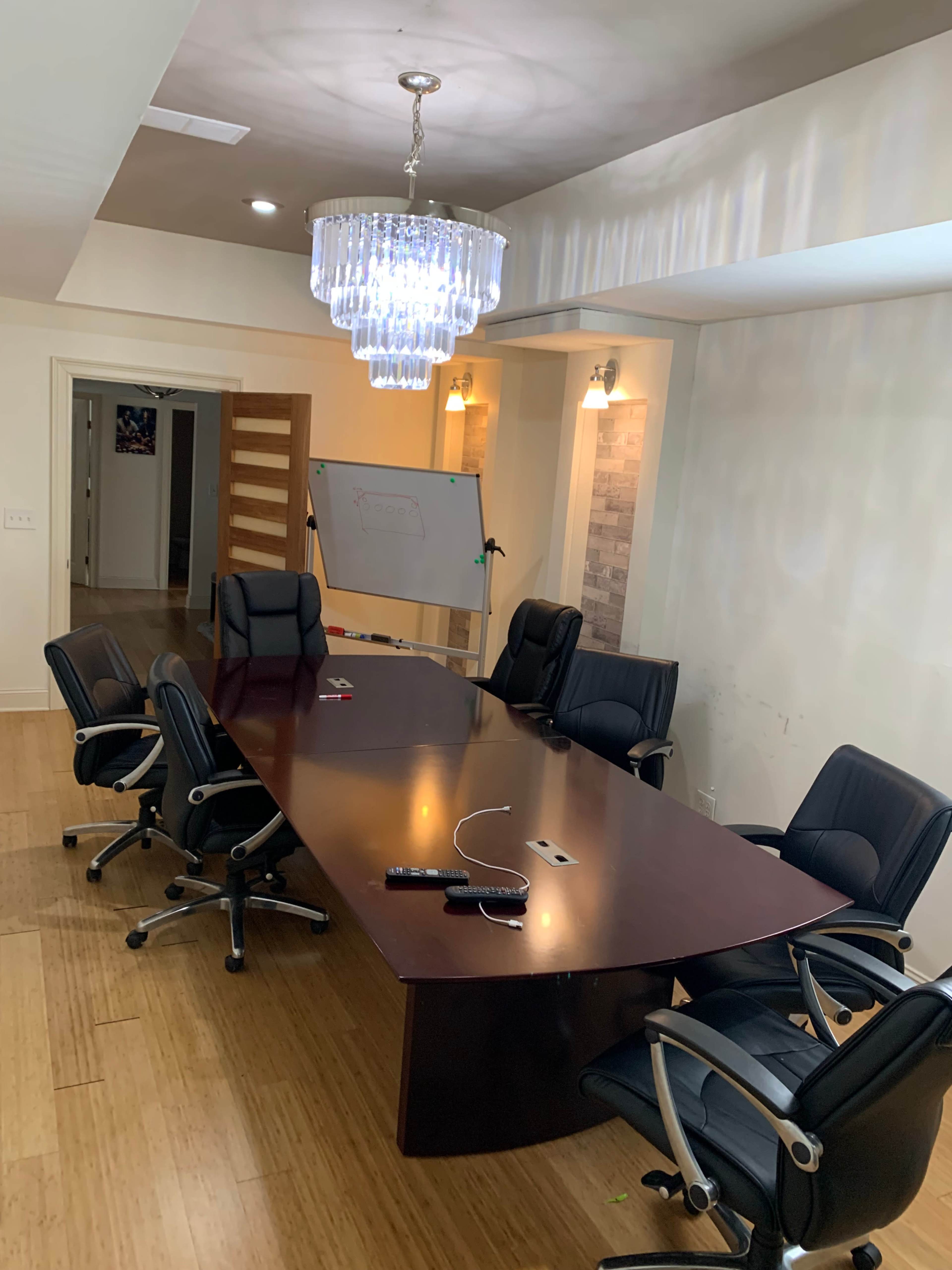 A large conference table surrounded by black ergonomic chairs is located in a well-lit meeting room with a decorative chandelier.