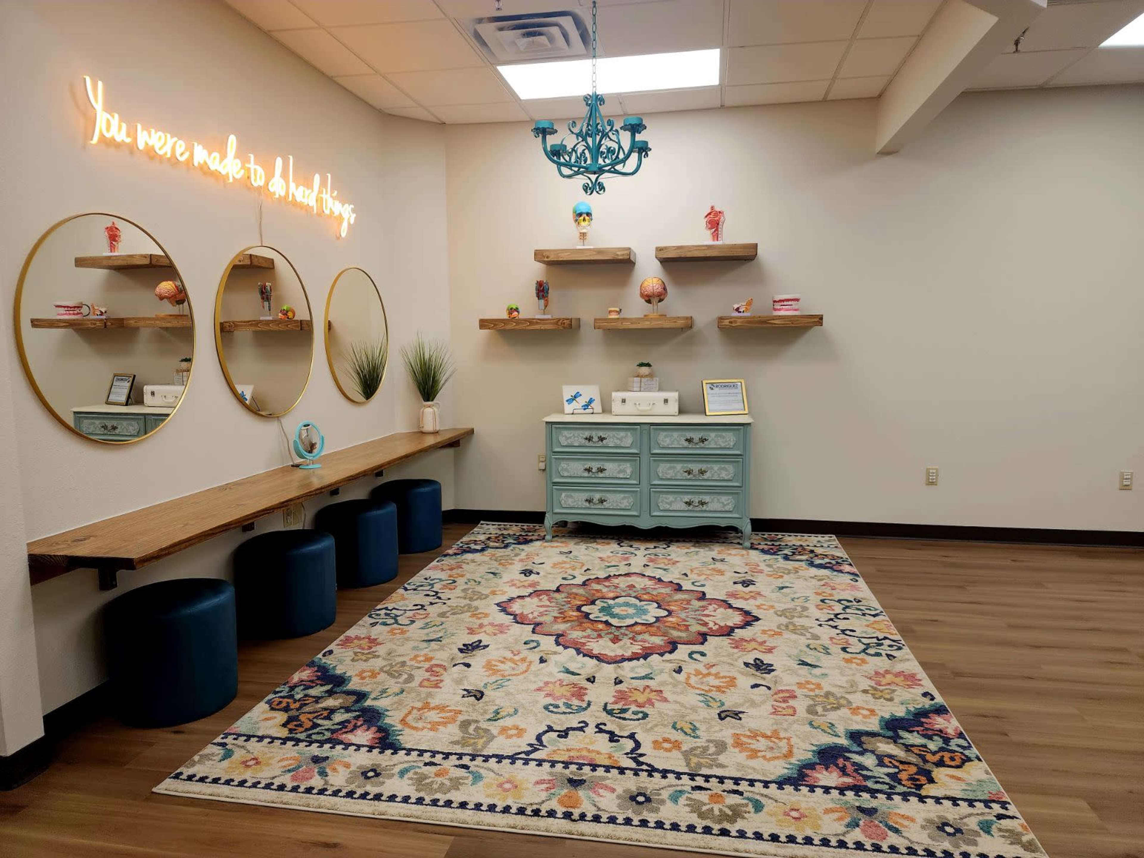 The image shows a well-lit room featuring circular mirrors on the wall, wooden shelves, a light blue dresser, and a patterned area rug, along with blue stools.