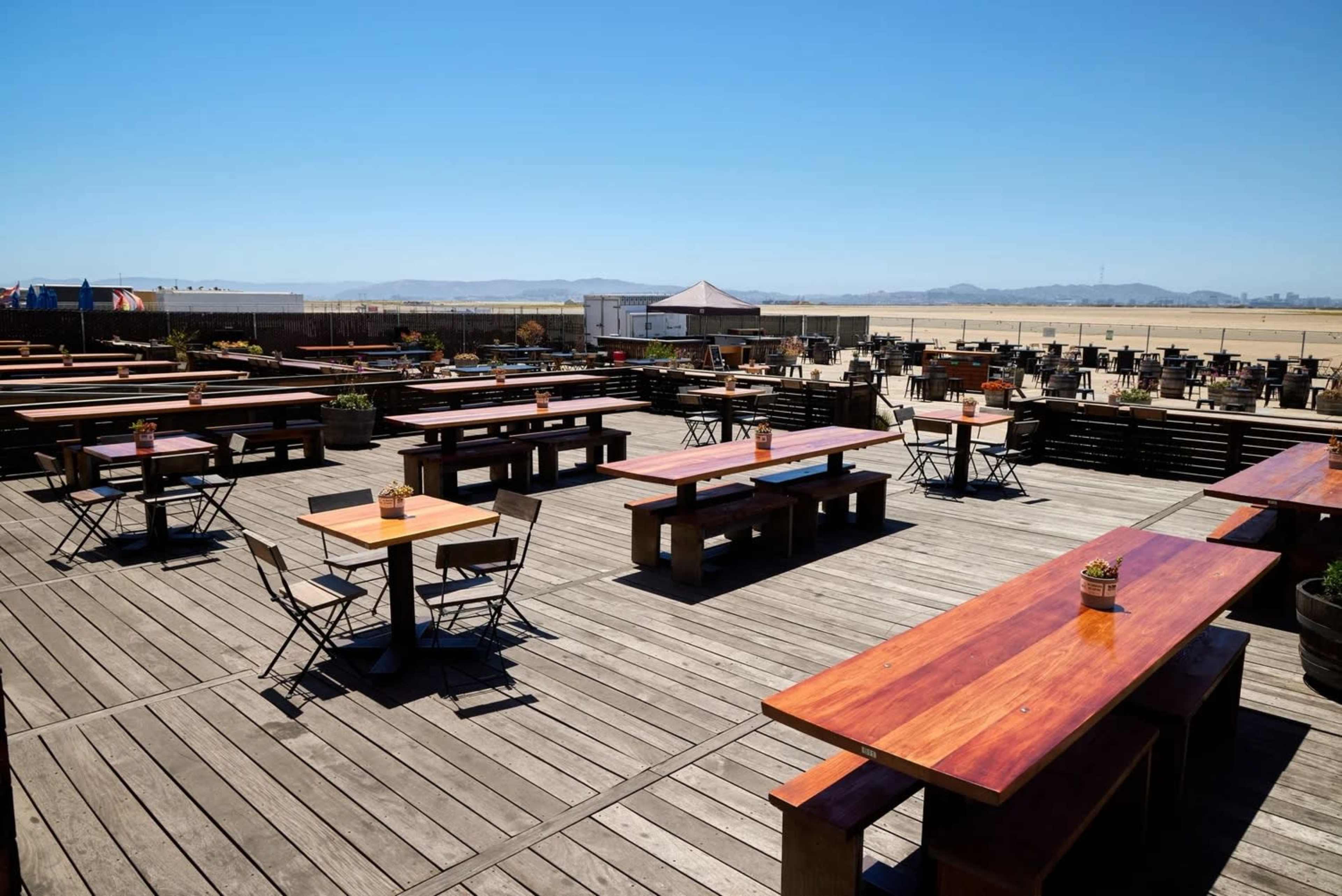 The outdoor dining area features numerous wooden tables and chairs situated on a wooden deck under a clear blue sky.