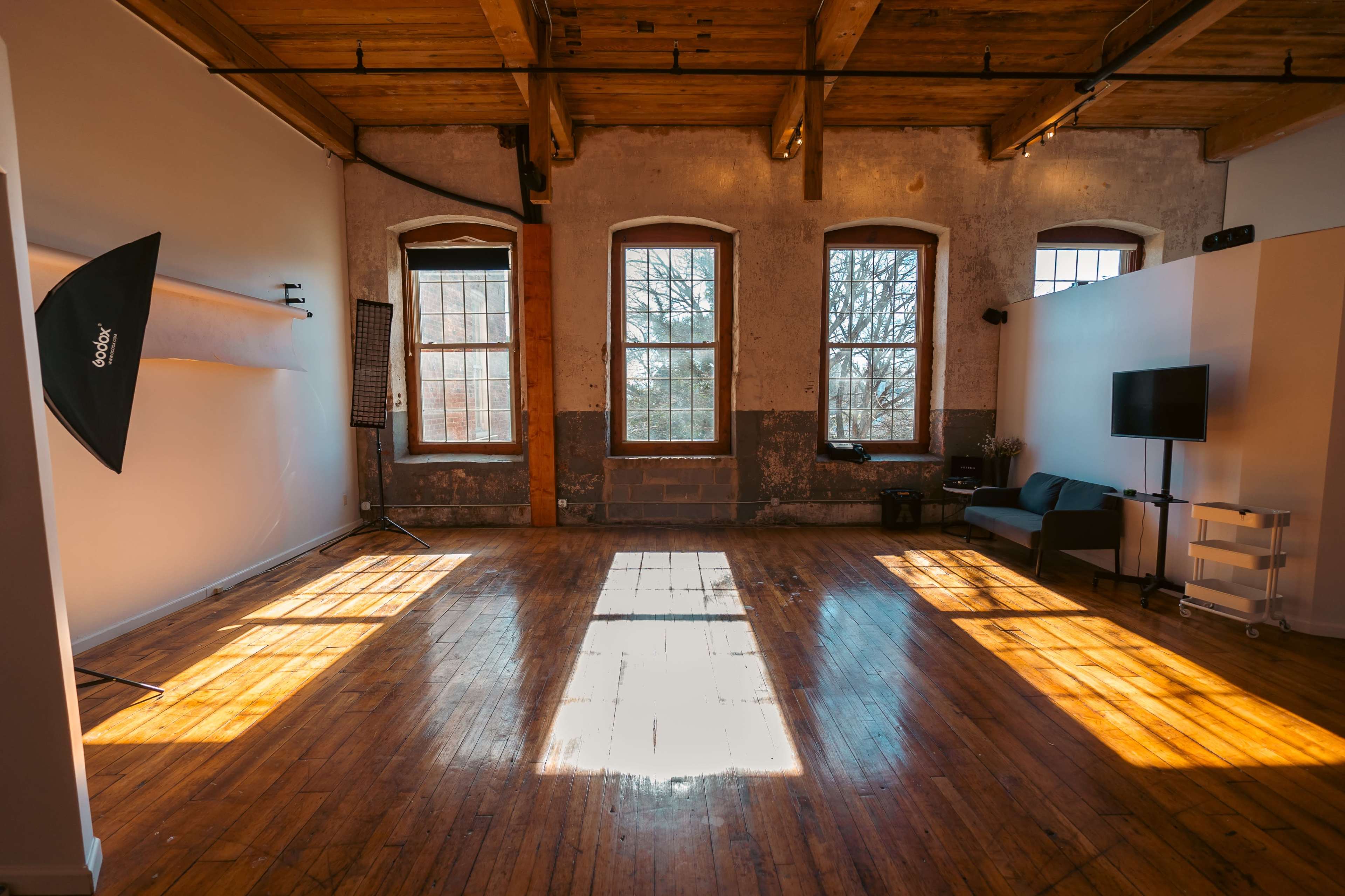 A spacious, well-lit studio with wooden floors, large windows, and a seating area.