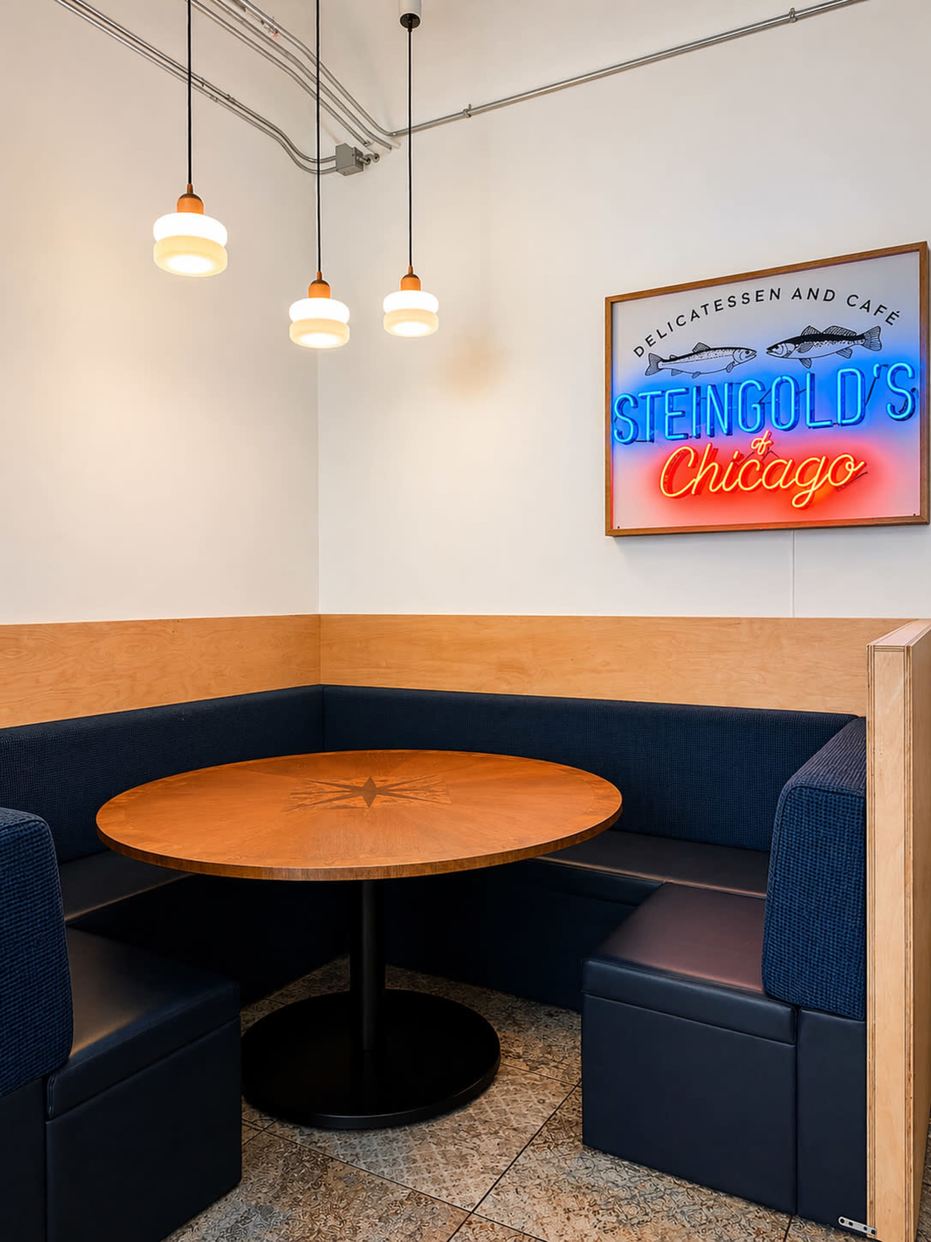 The image shows a corner seating area in a cafe, featuring a round wooden table surrounded by upholstered benches and illuminated by pendant lights, with a neon sign on the wall.