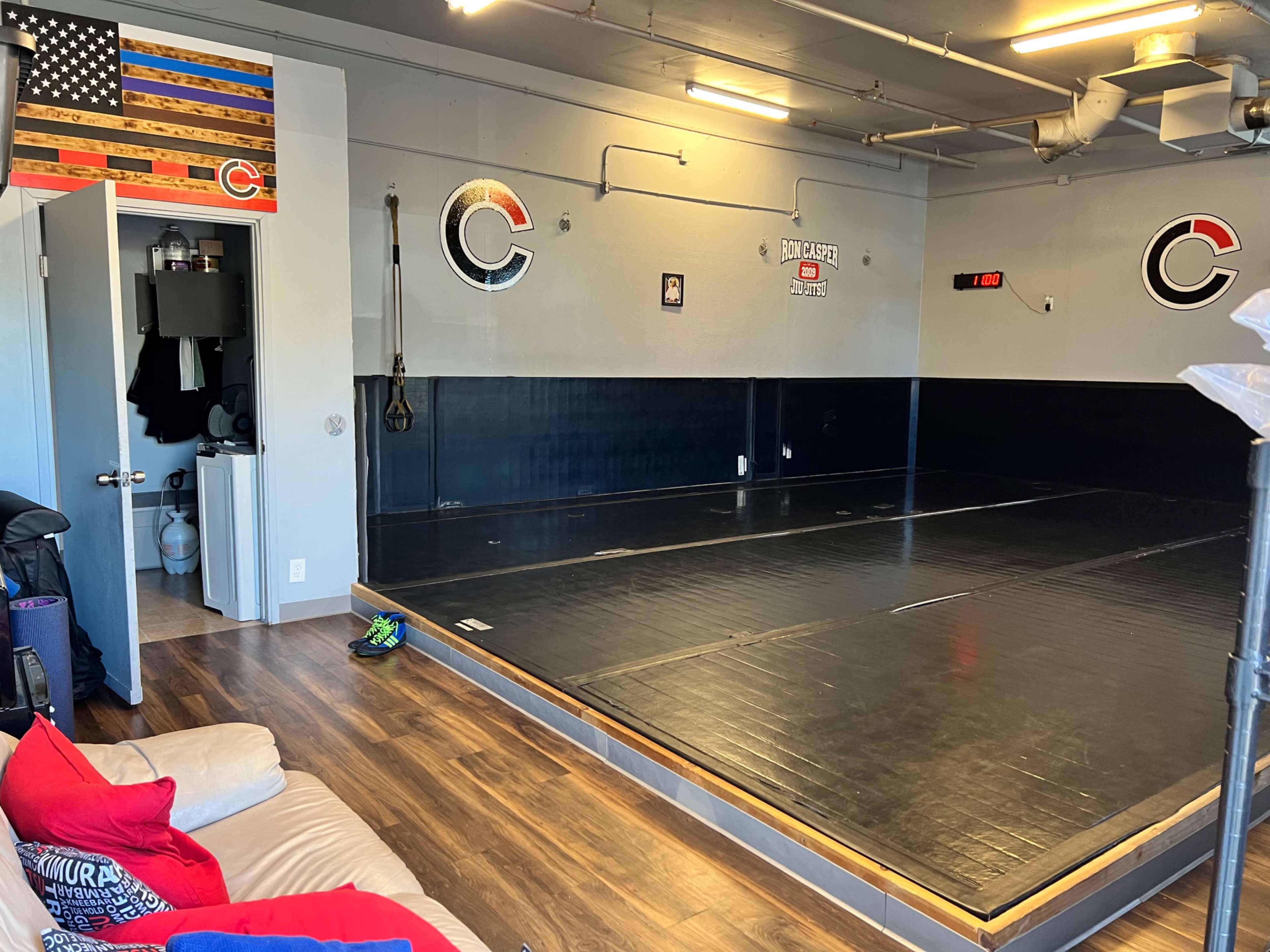 The image shows a training room with a large, black mat space, an American flag on the wall, and a doorway leading to a restroom area.