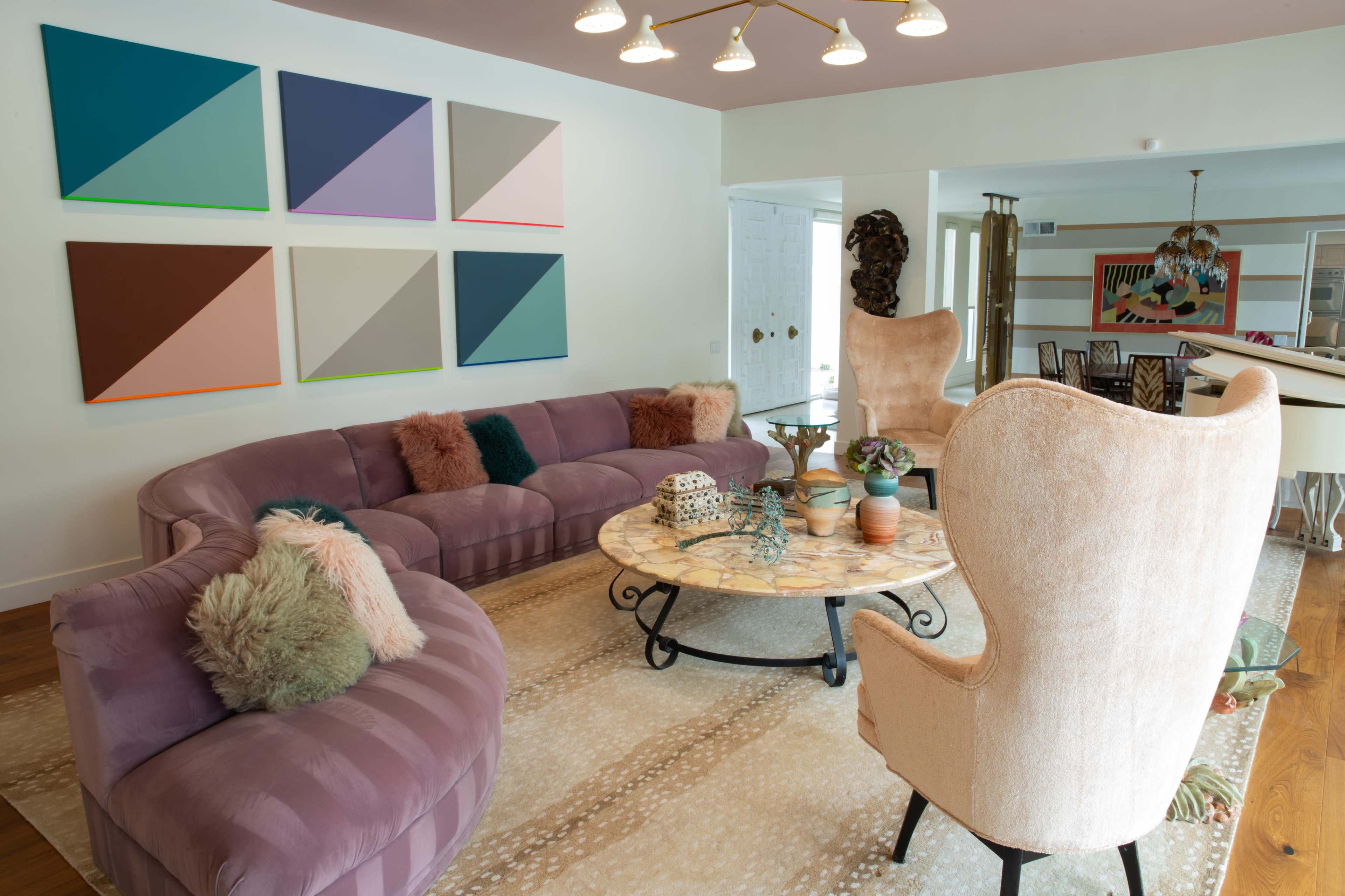The room features a curved purple sofa, a round coffee table with decorative items, and two large armchairs, all arranged around a patterned area rug under a chandelier.