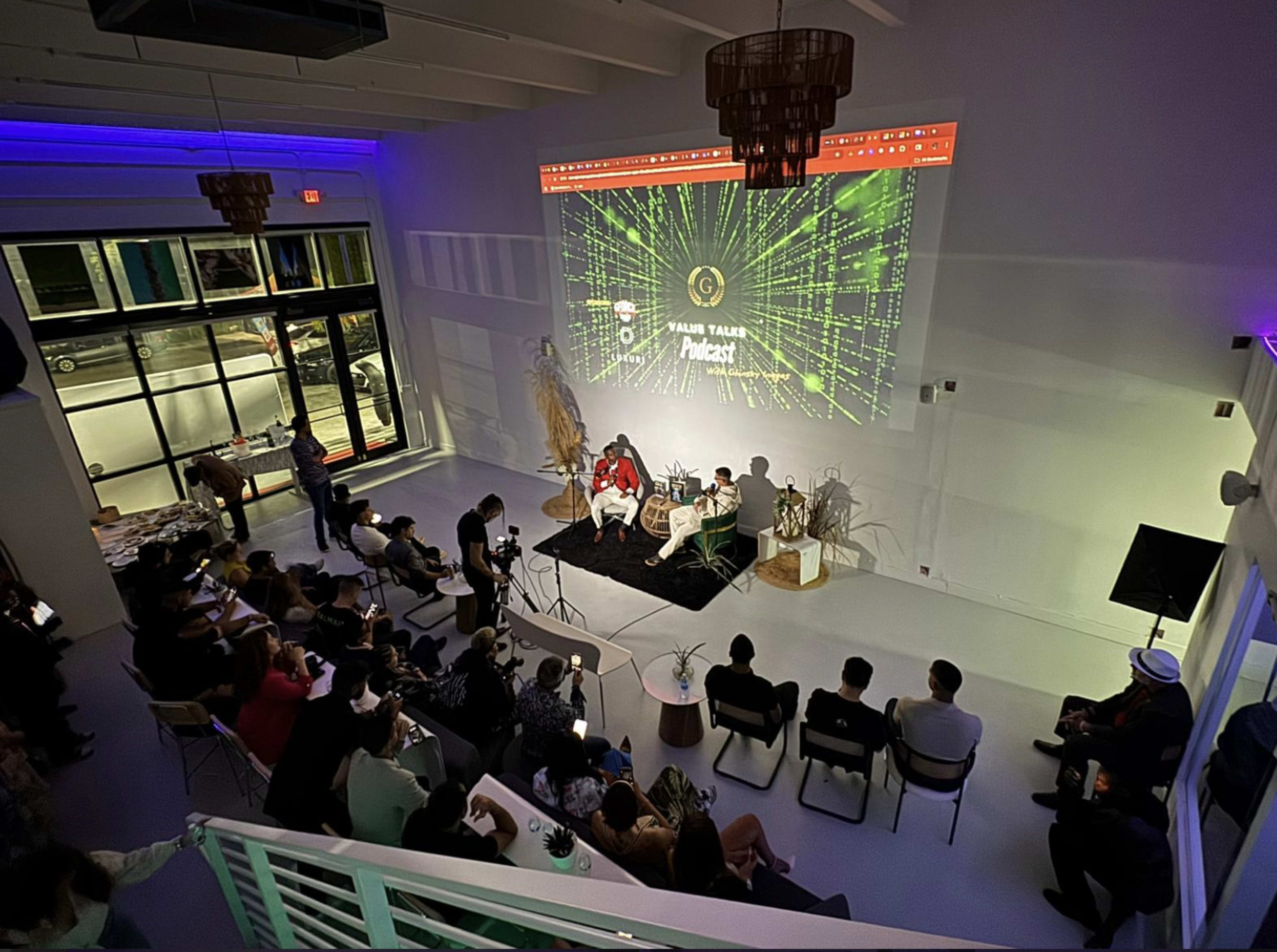 A crowd sits in a well-lit modern venue, watching a podcast discussion featuring two speakers on a stage with a large digital backdrop.