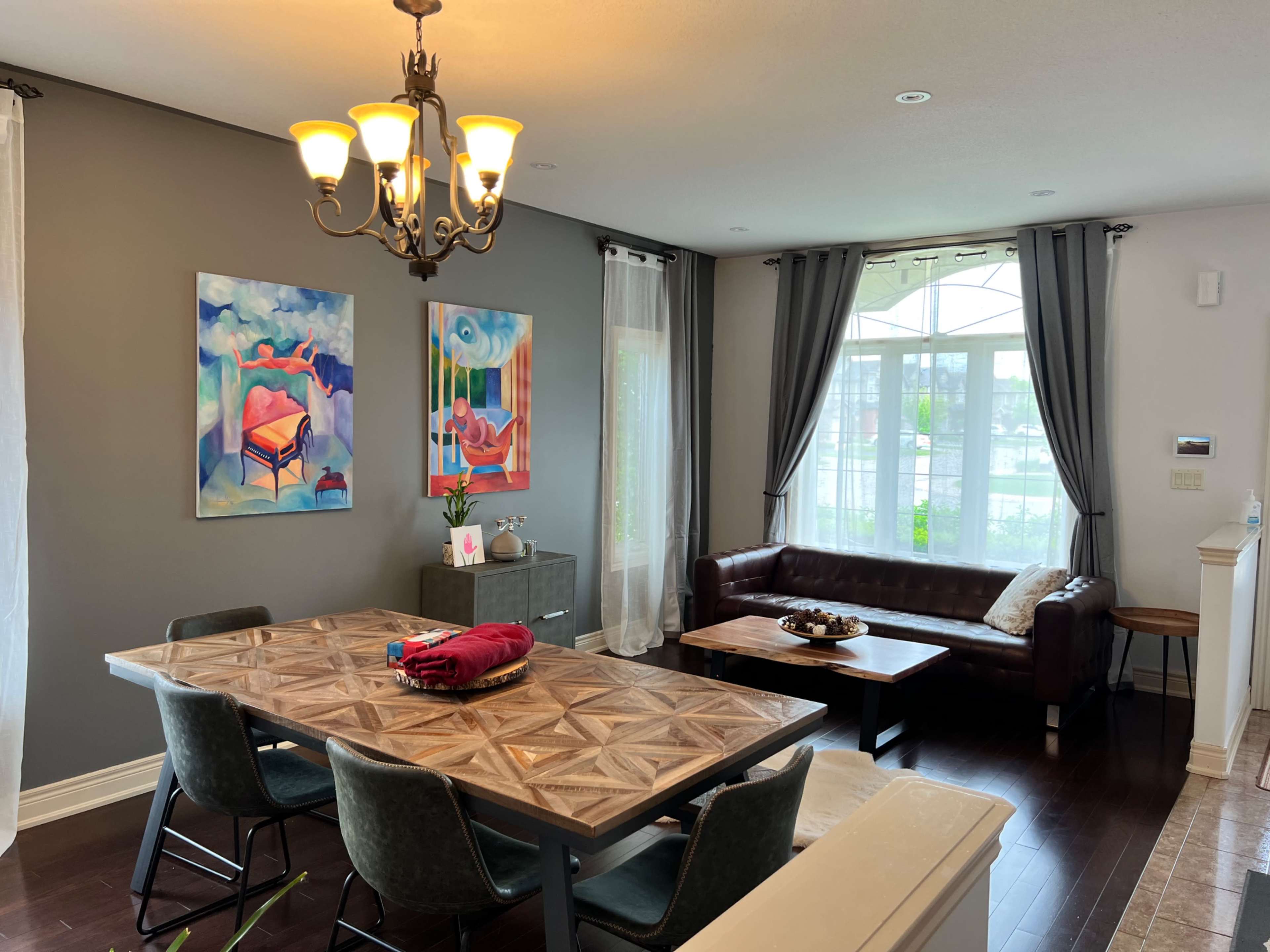 The image shows a dining area featuring a wooden table set with chairs, a couch, artistic wall paintings, and large windows providing natural light.