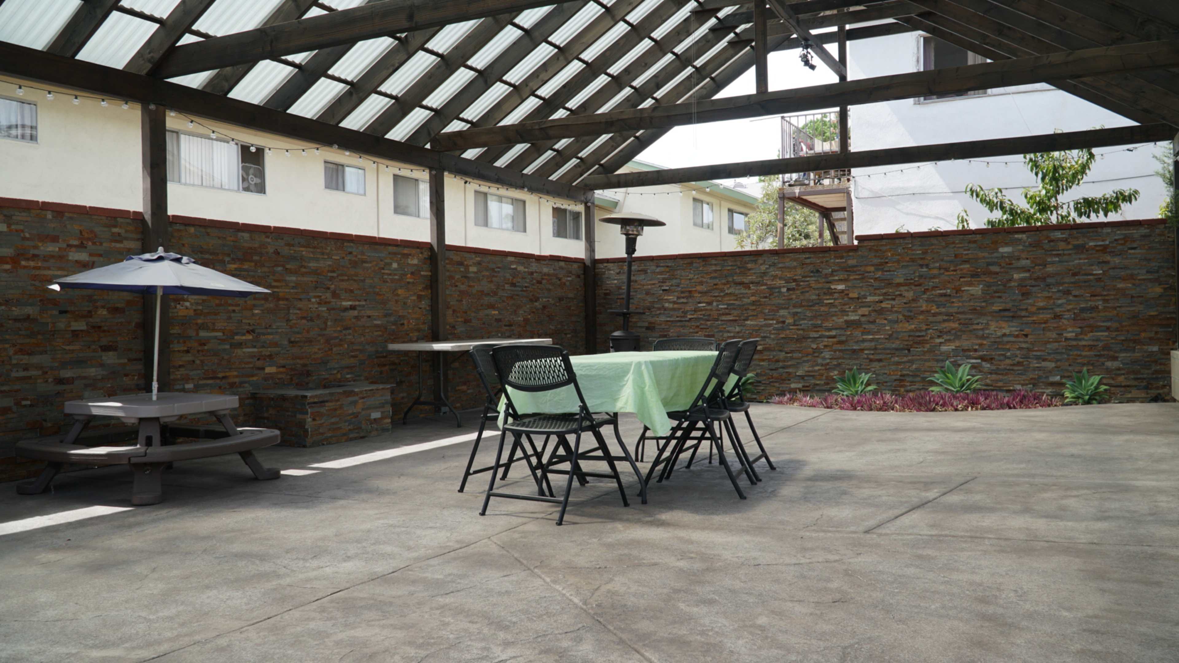 The image shows a spacious outdoor patio area with a green tablecloth on a large table, surrounded by chairs, and a shaded picnic table nearby.