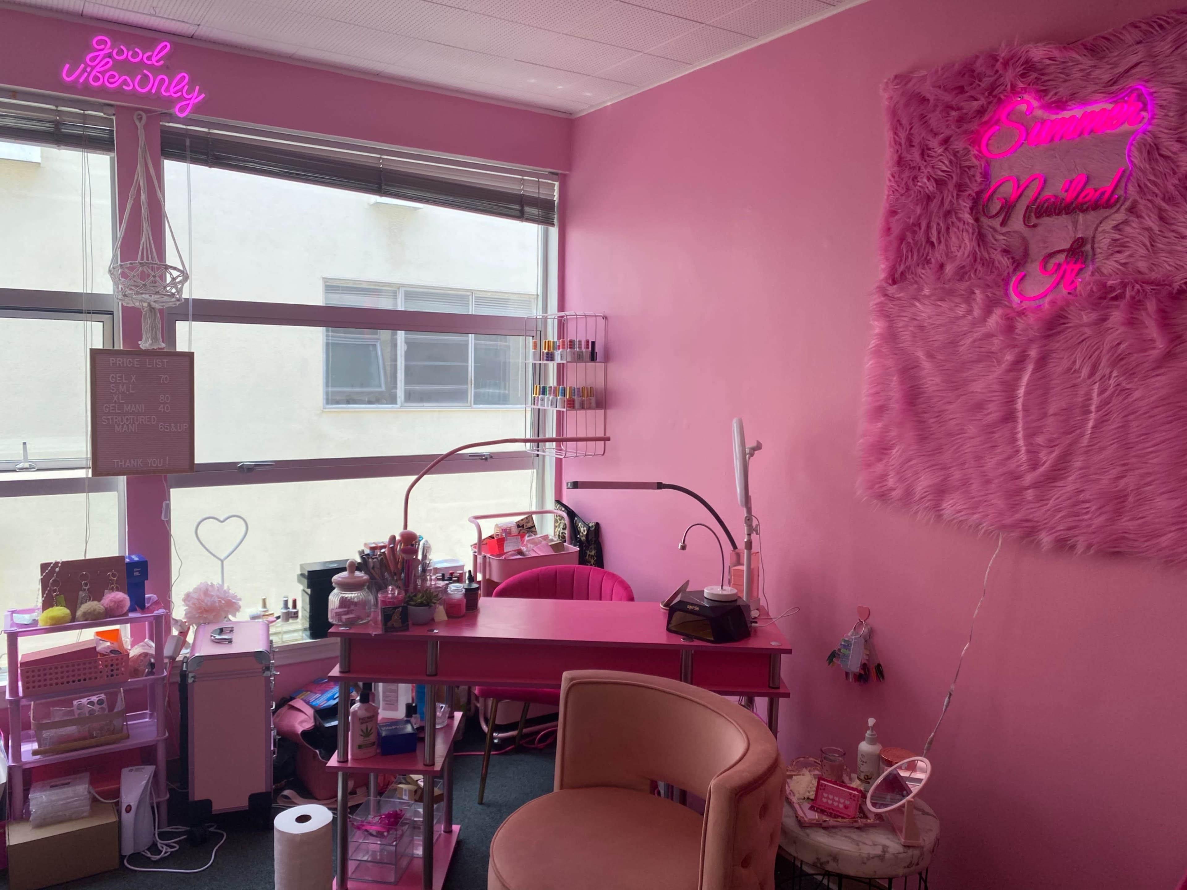 A brightly colored pink room features a nail station with a lamp, storage for nail products, and neon signs on the walls.