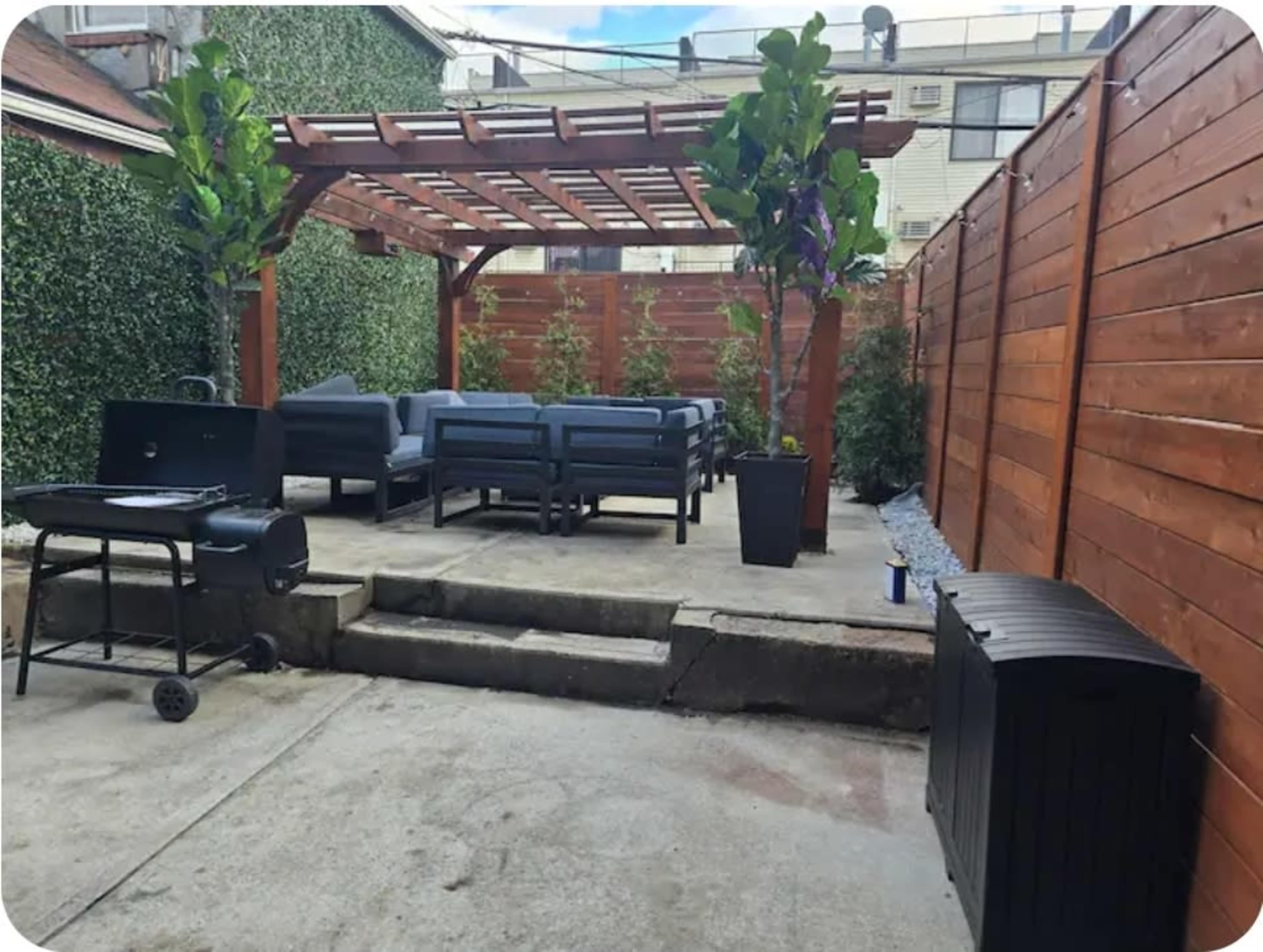 The image shows a patio area with a wooden pergola, seating arrangements, and a grill, surrounded by tall plants and wooden fencing.