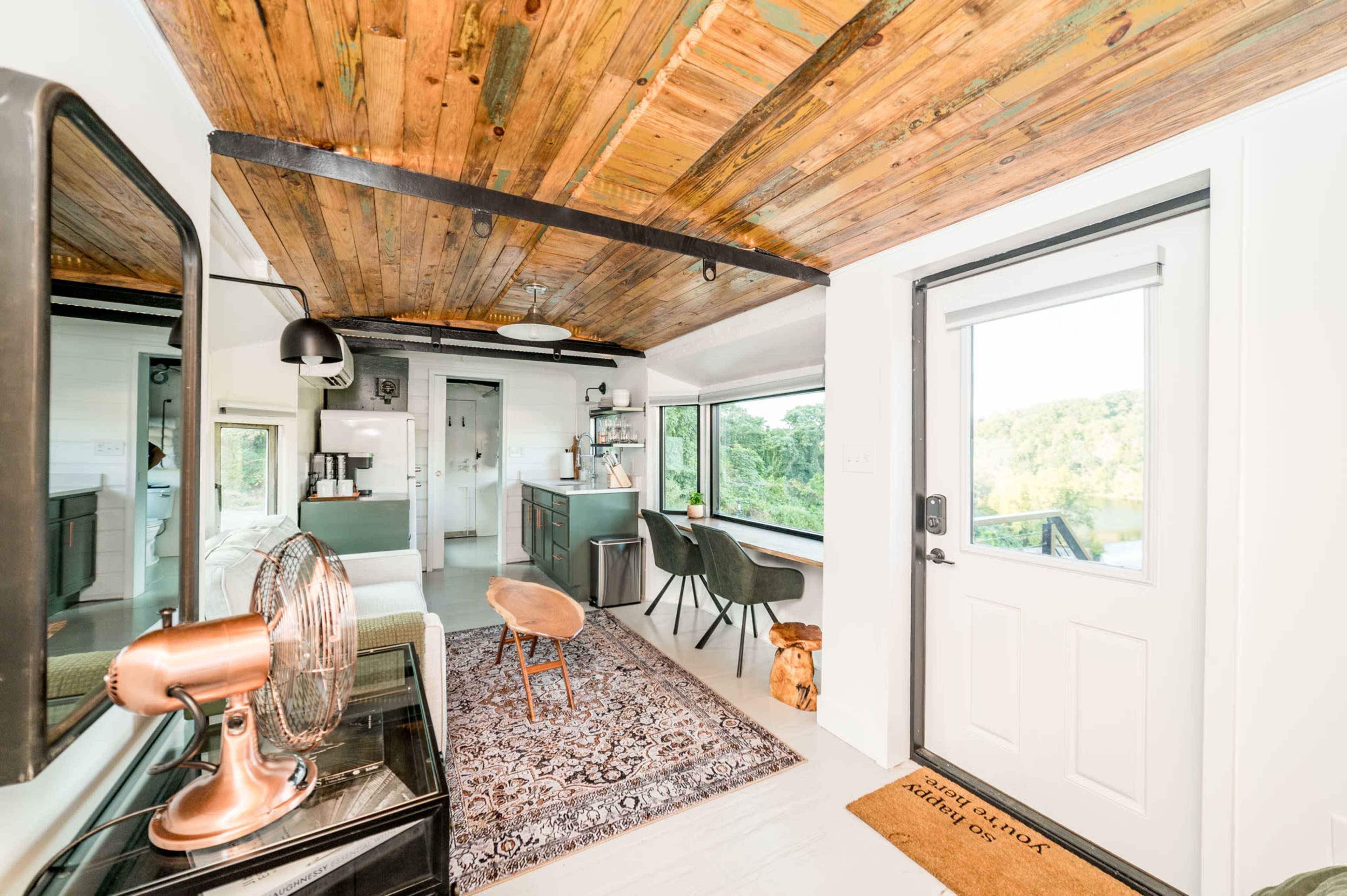 A small, modern cabin interior featuring wooden beams, a stylish living area with a rug, a kitchen area, and a front door leading to an outdoor view.