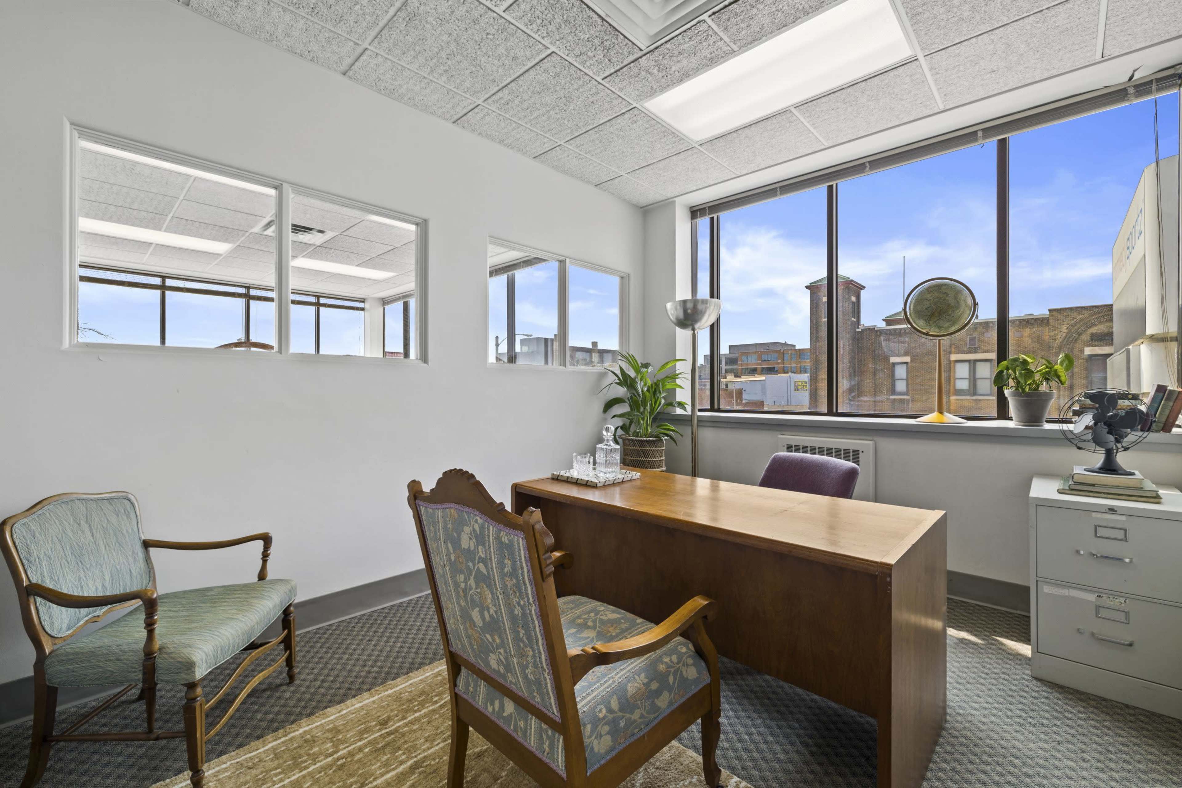 The image shows a bright office space featuring a wooden desk, an armchair, and a large window with a view of a cityscape.