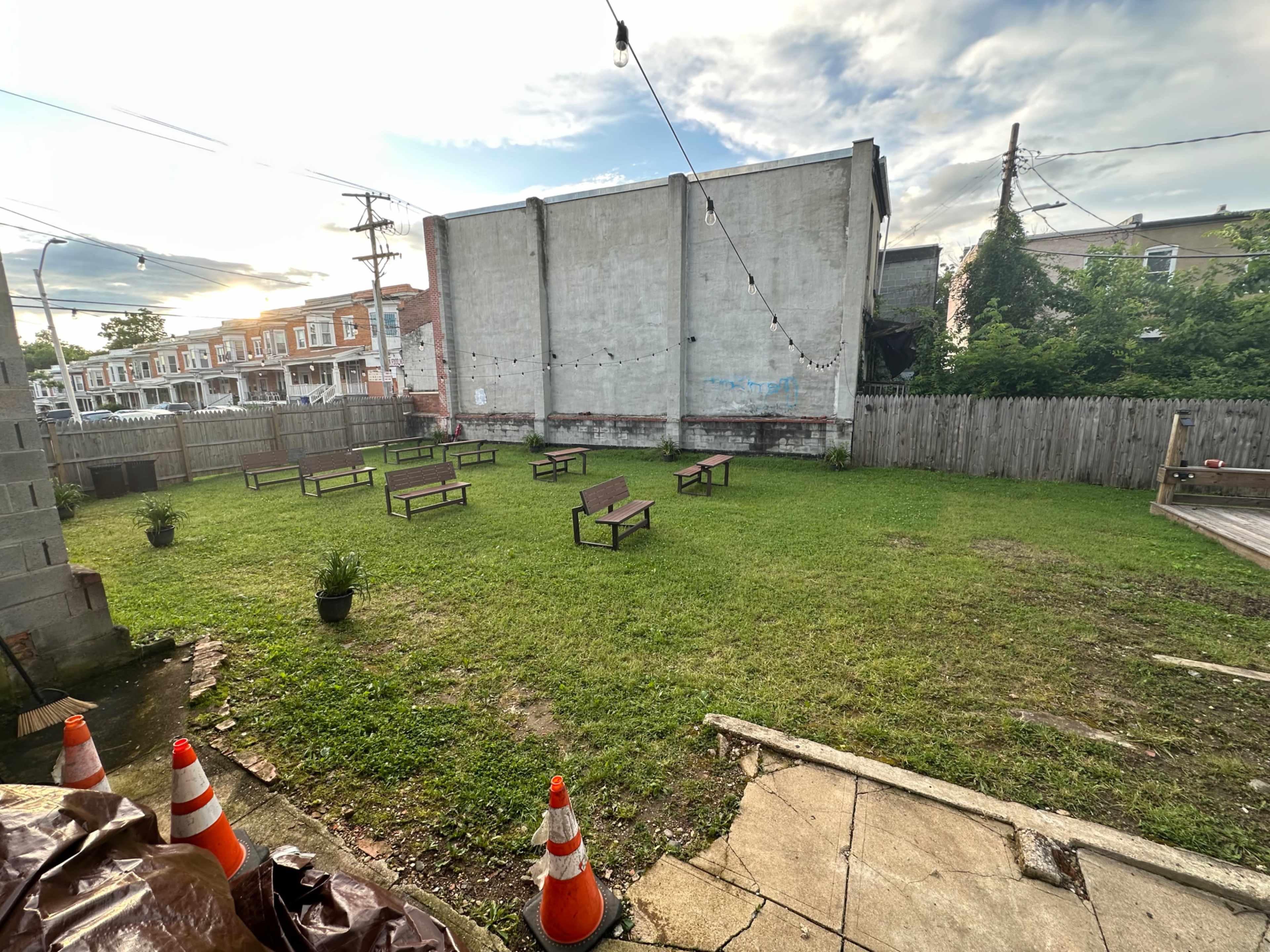 The image shows a grassy outdoor area with several wooden benches arranged in rows, surrounded by a wooden fence and a concrete walkway.