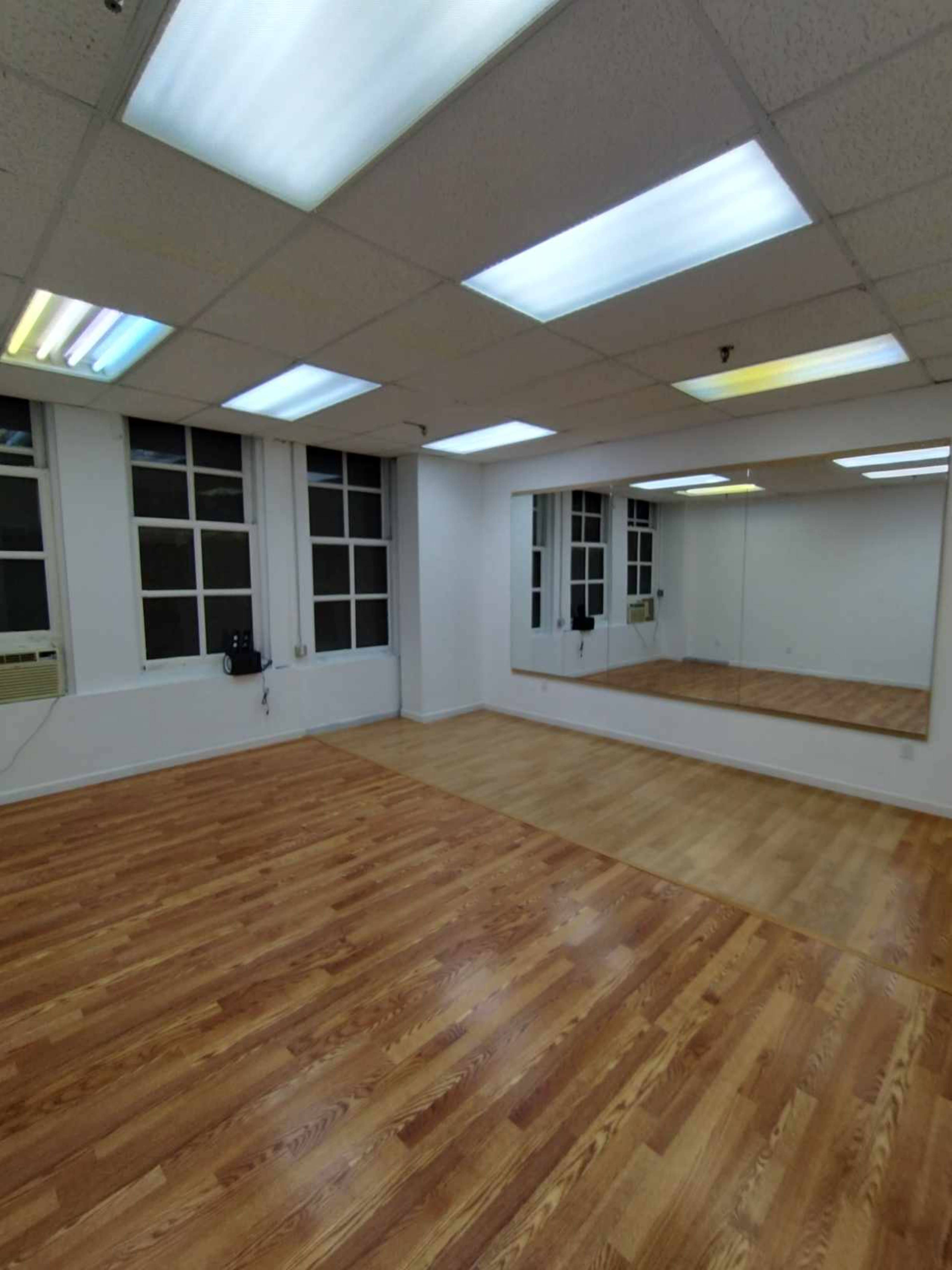 The image shows a spacious room with hardwood flooring, large windows, and a mirror on one wall.