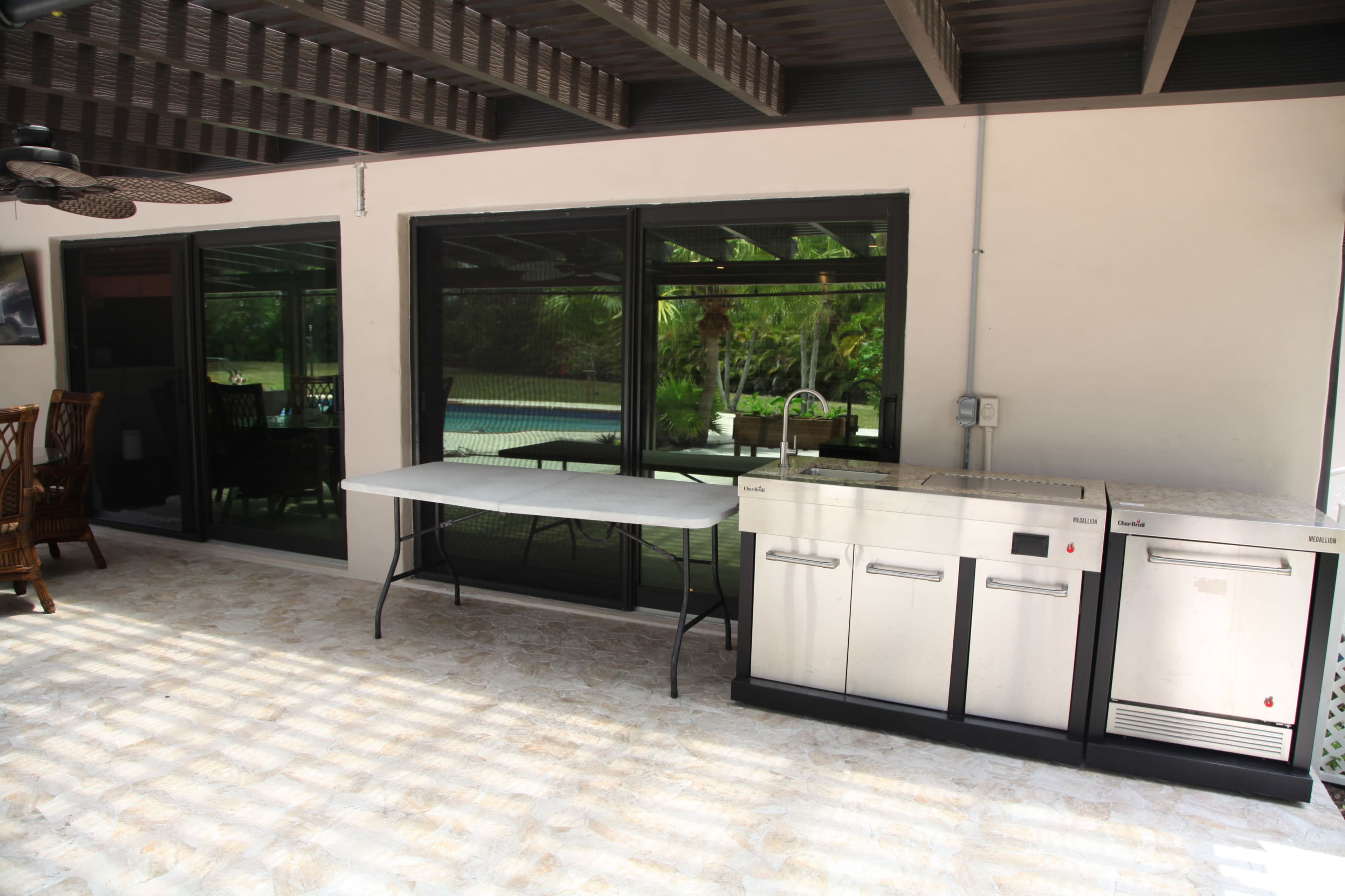 An outdoor kitchen area featuring a stainless steel grill and refrigerator, with a table set beside large windows overlooking a swimming pool.